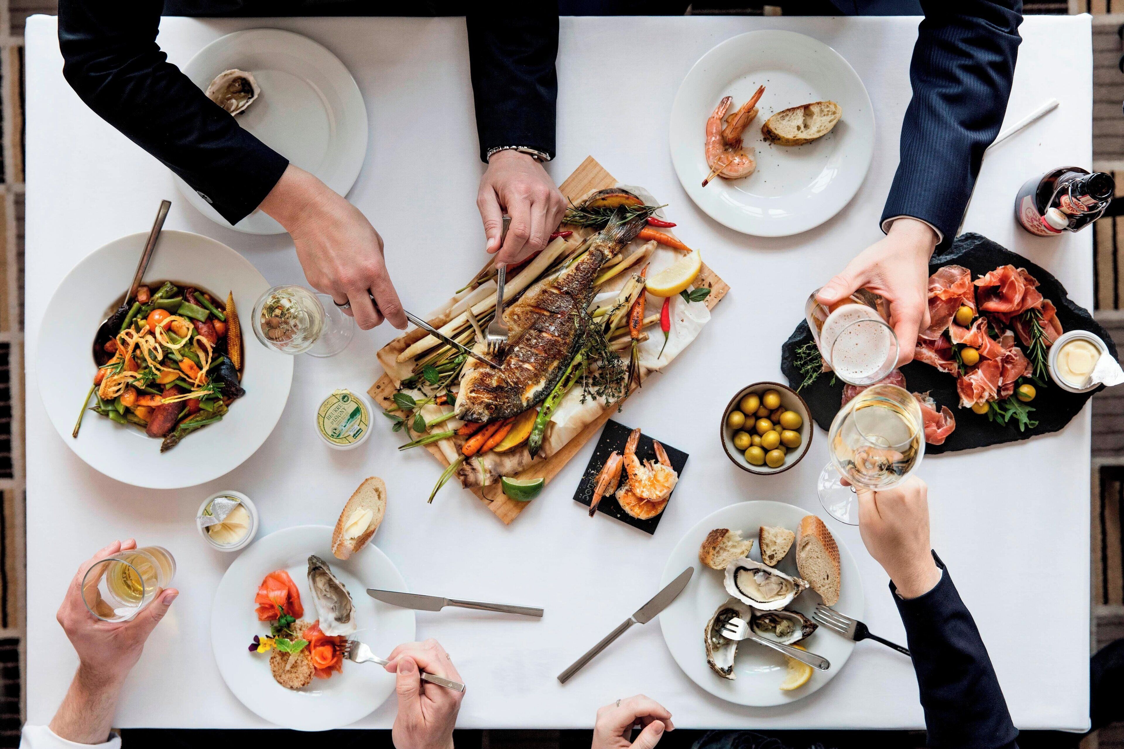 A group of people dining at a table with various dishes, including seafood, bread, and a fish centerpiece.