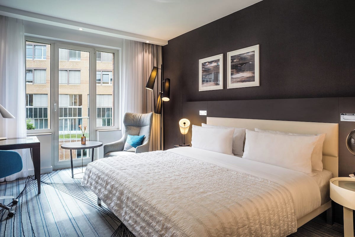 A hotel room with a large bed, a desk, a chair, a lamp, and a view of the outside.