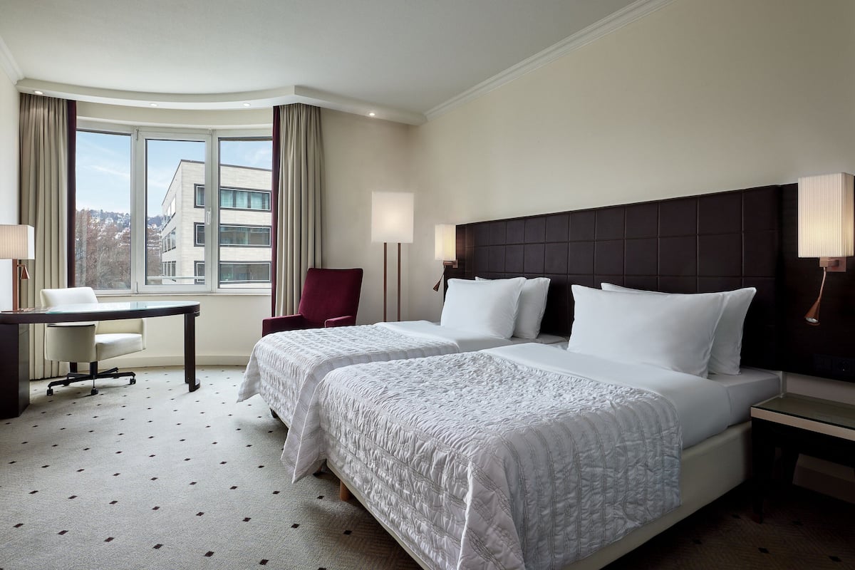 A hotel room with two beds, a desk, a chair, and a large window offering a city view.
