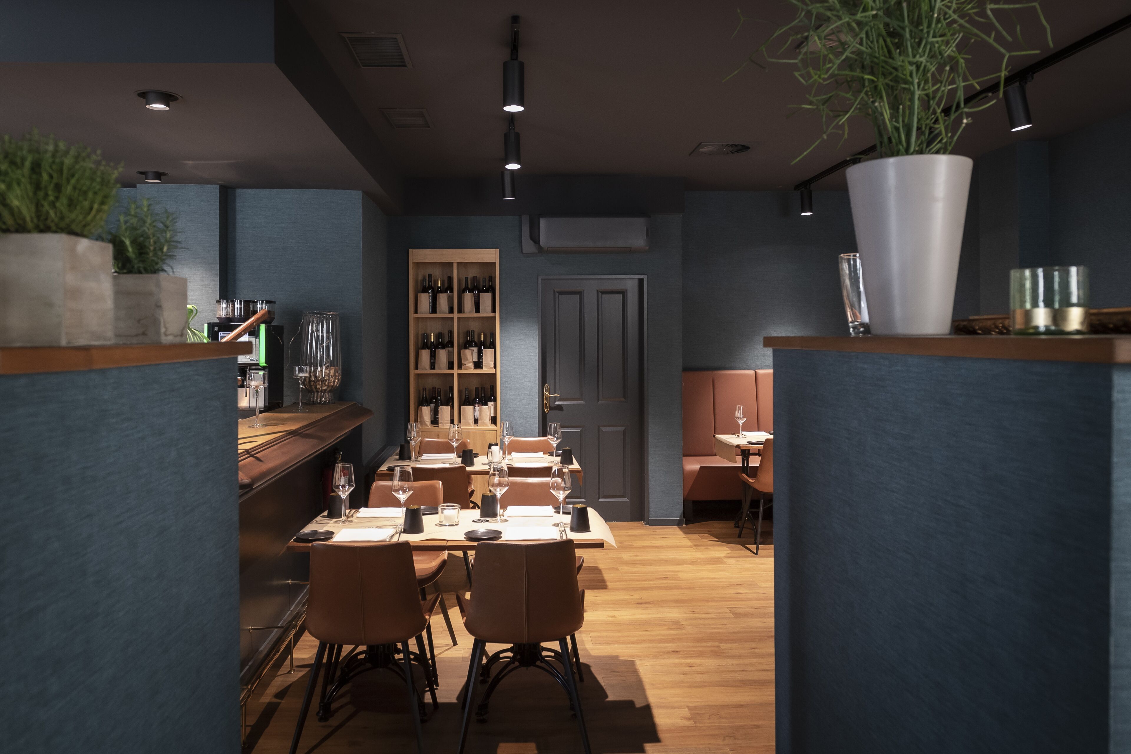 Un comedor moderno con mesas y sillas de madera, una barra con planta en maceta y una estantería para vinos al fondo.