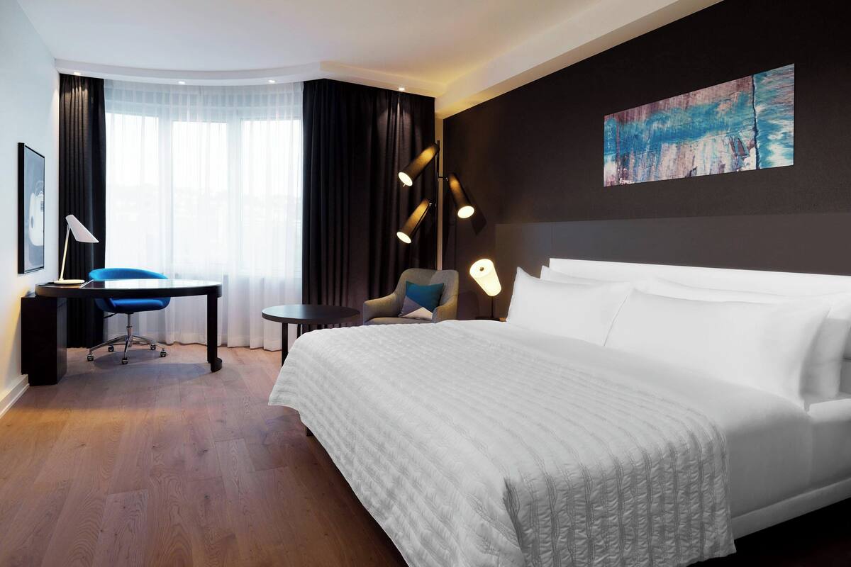 A hotel room with a large bed, a desk, a chair, and a painting on the wall.