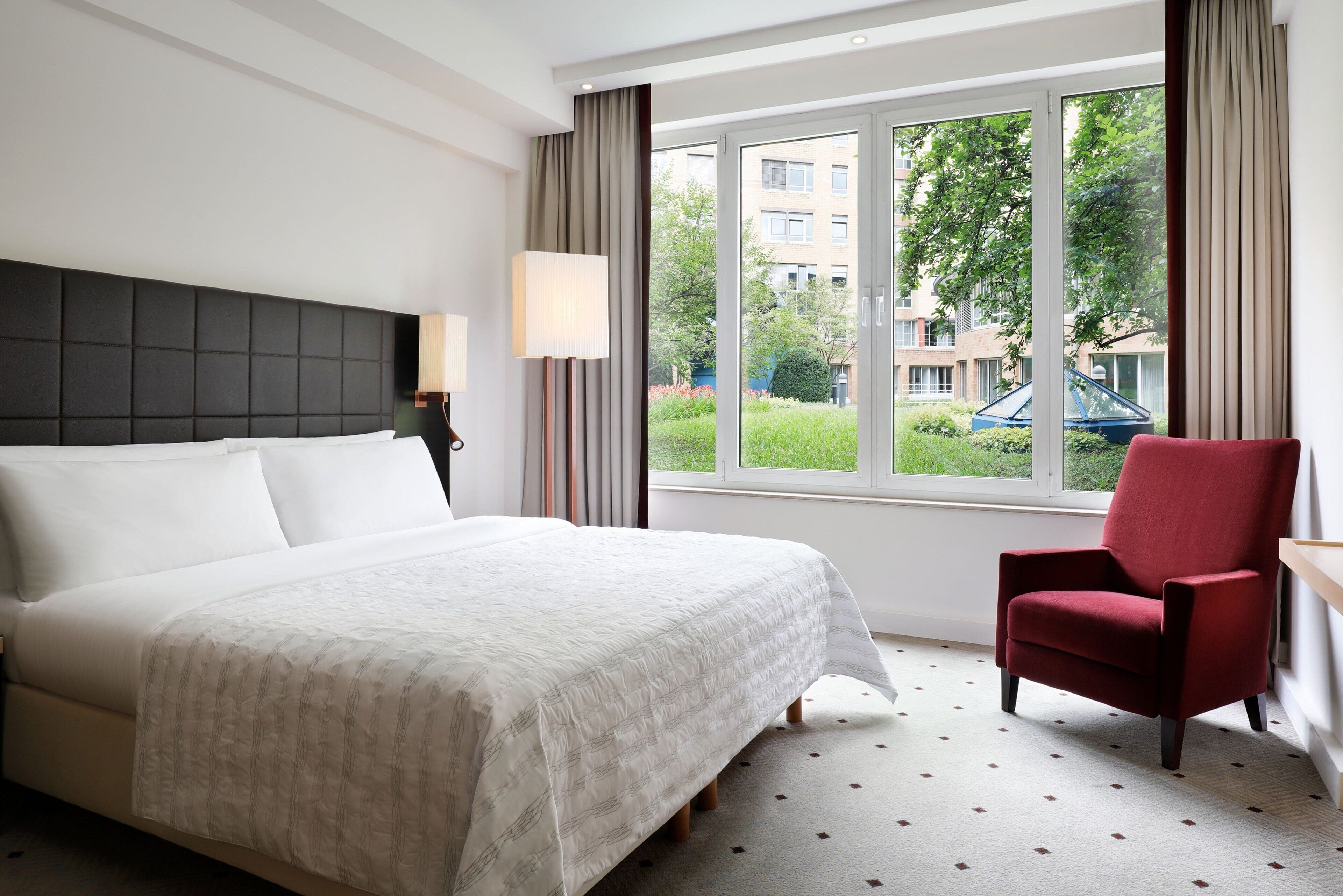 A hotel room with a large bed, a red armchair, a window with a view of buildings and greenery, and a lamp on the nightstand.