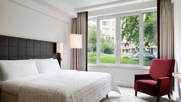A hotel room with a large bed, a red armchair, a window with a view of buildings and greenery, and a lamp on the nightstand.