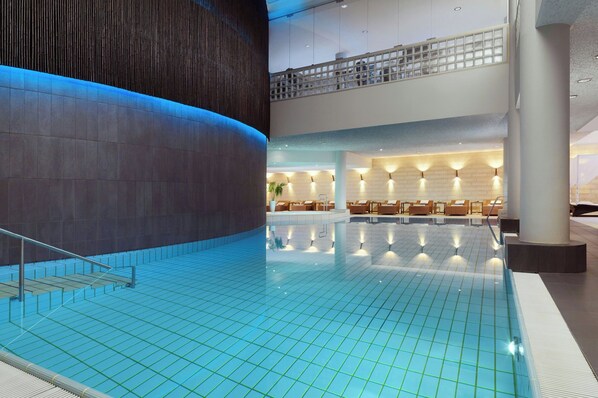 A modern indoor swimming pool with a high ceiling and a seating area with chairs and tables.