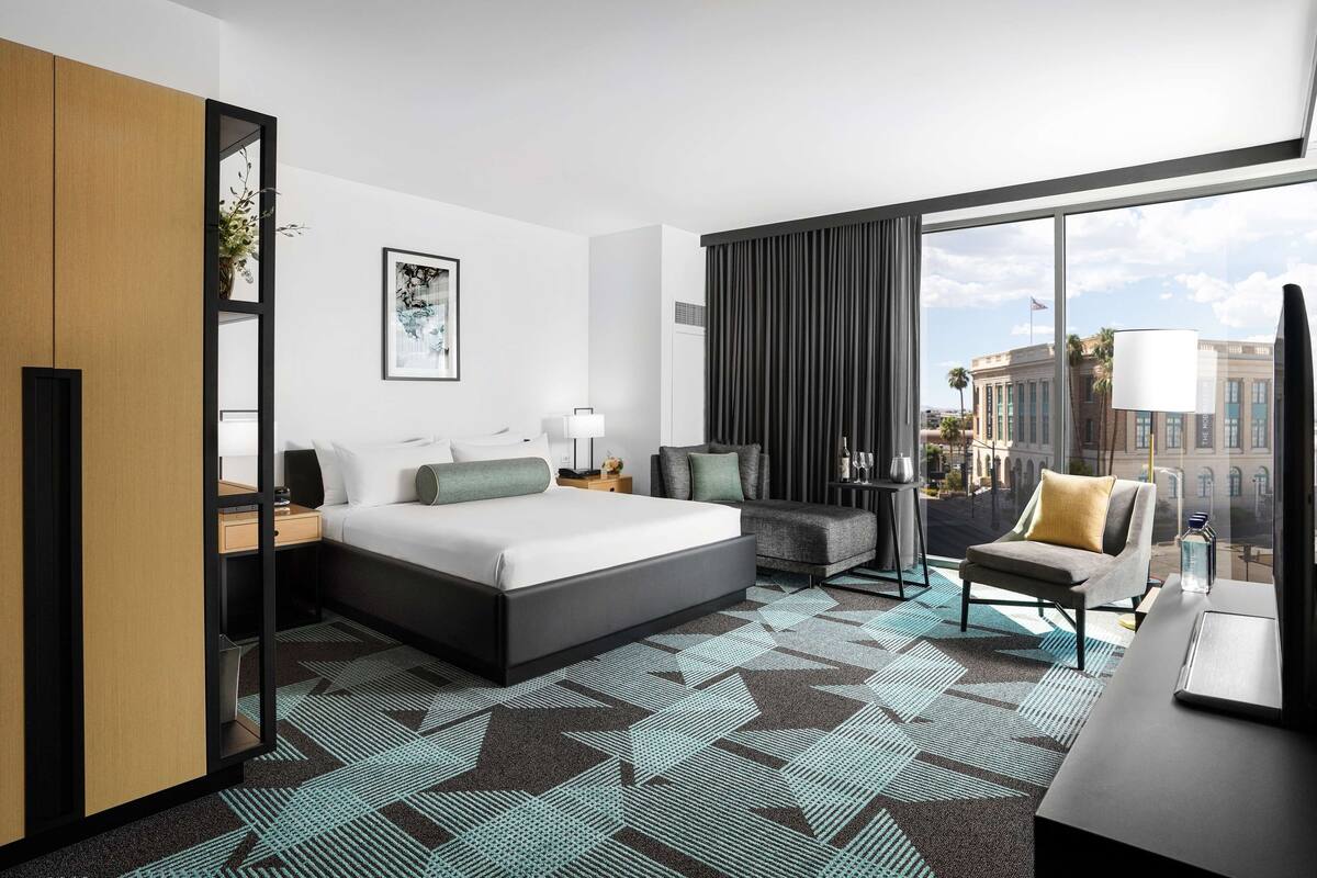 Quarto de hotel com uma cama grande, uma escrivaninha, uma cadeira, uma janela ampla com vista para edifícios e um tapete estampado.