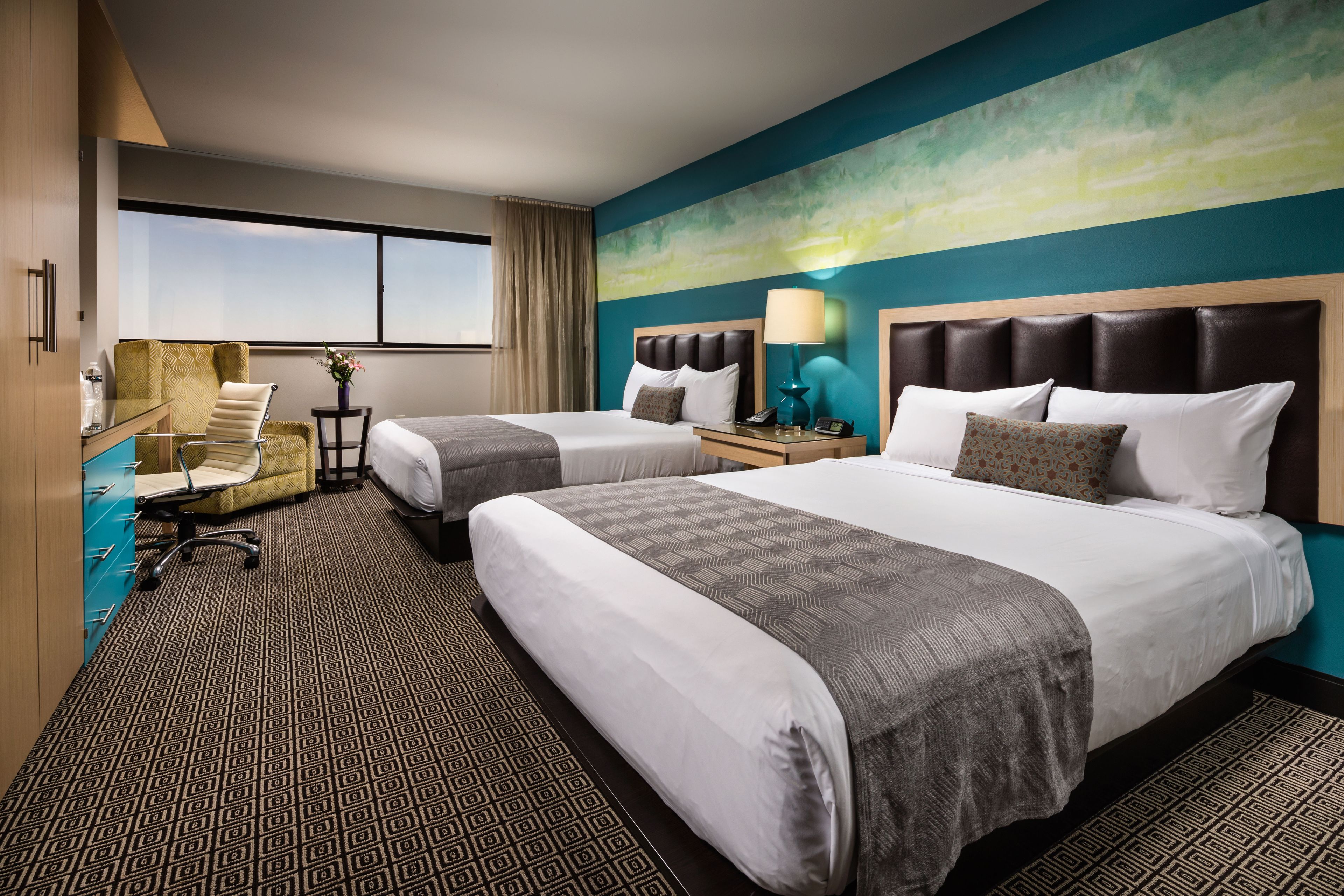 A hotel room with two beds, a desk, a chair, a window, and a painting on the wall.