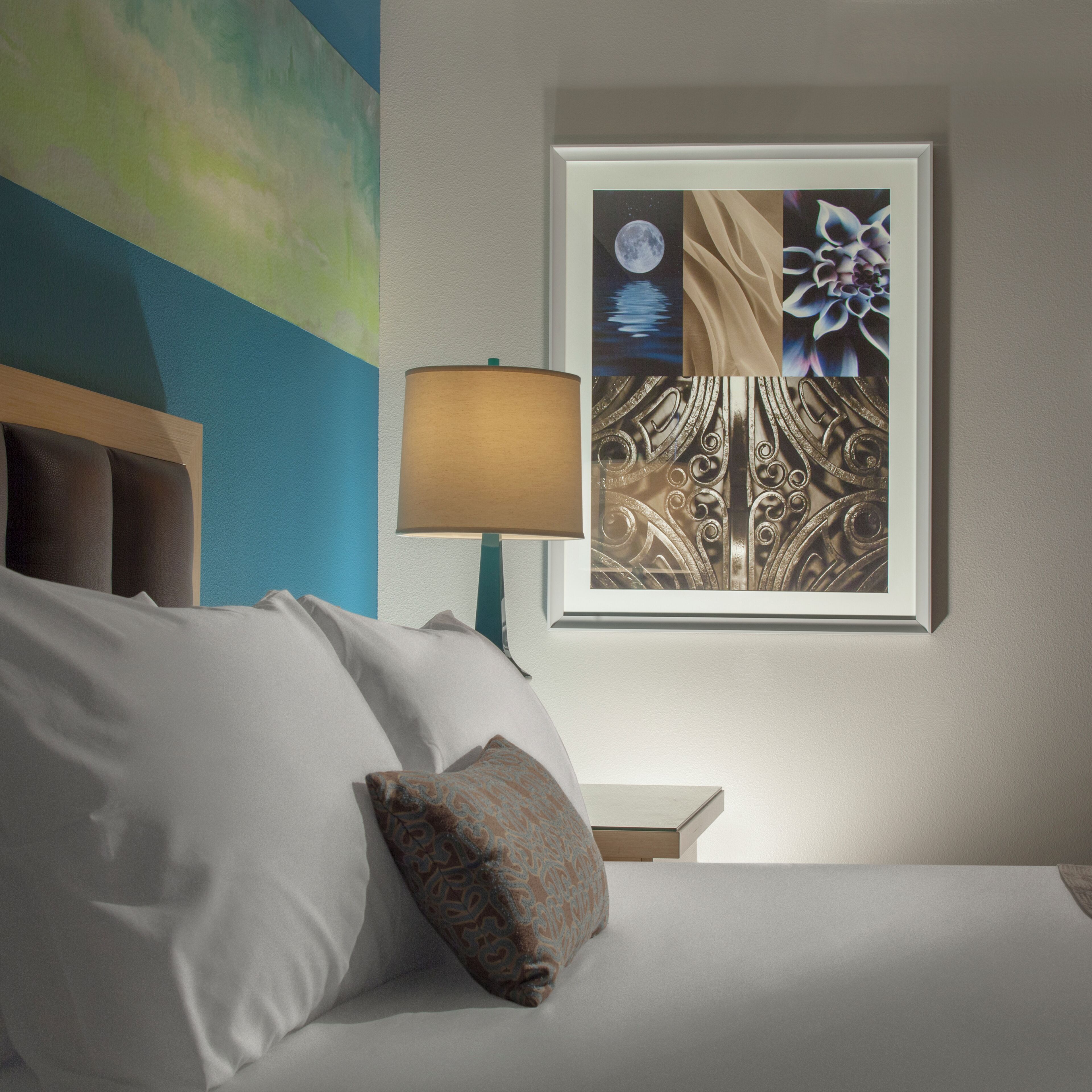 Een net opgemaakt bed met witte lakens en een enkel decoratief kussen, een nachtkastje met een lamp, en een lijstwerk met een afbeelding van een nachtelijke scène met een maan en bloemistische details aan de muur.