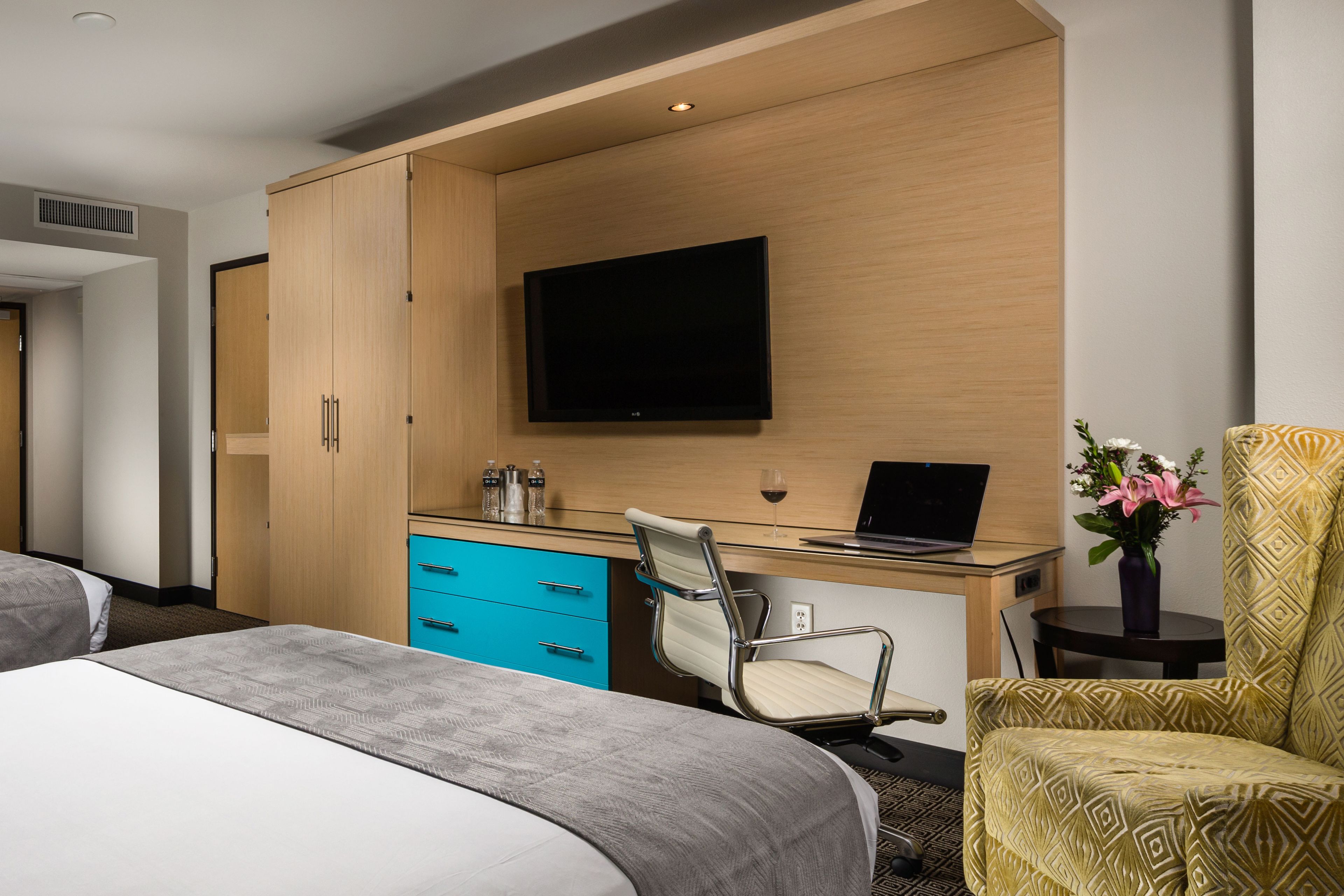 A hotel room with a bed, a desk with a laptop, a chair, a TV mounted on the wall, and a small table with a vase of flowers.