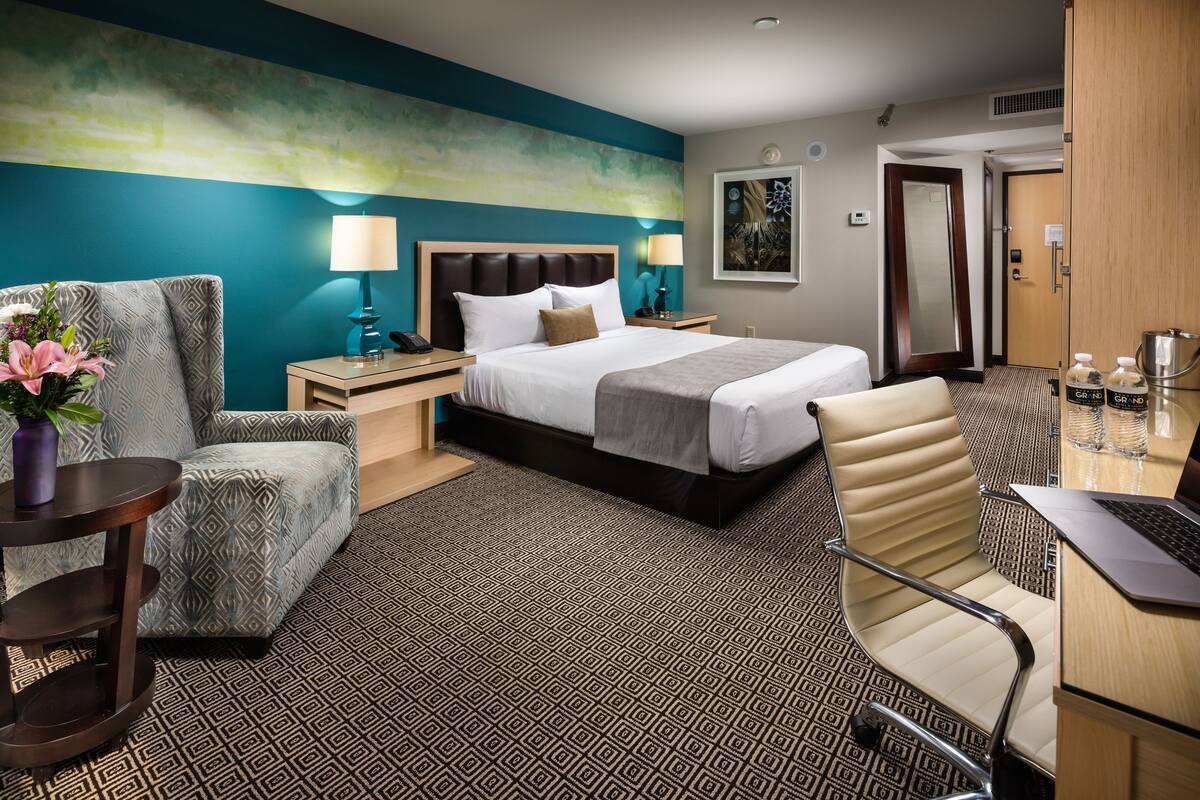 Quarto de hotel com uma cama grande, uma cadeira, escrivaninha com um laptop, mesa de cabeceira com abajur e porta do banheiro.