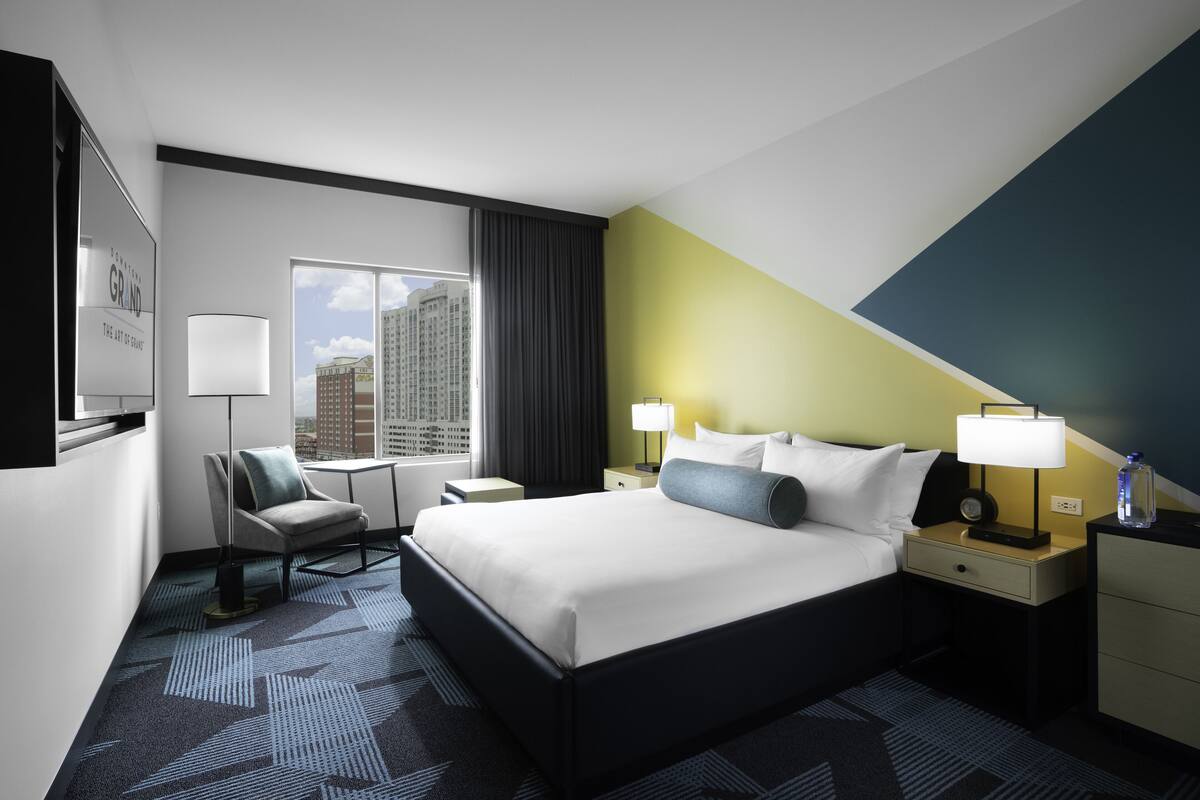 Quarto de hotel com cama, duas mesinhas de cabeceira, uma cadeira, escrivaninha, luminária, televisão e janela com vista para a cidade.