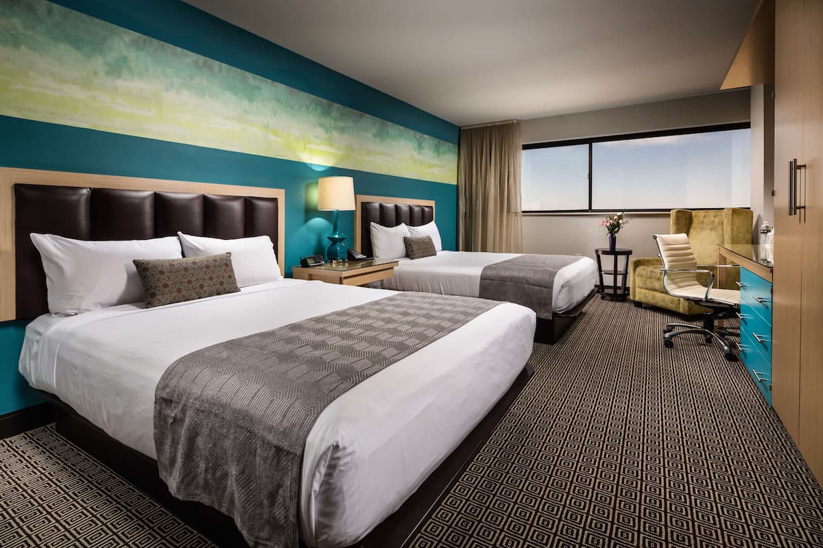 Quarto de hotel com duas camas, uma escrivaninha, uma cadeira e uma janela grande.