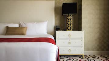 Un lit bien fait, avec du linge de lit blanc et un élément décoratif rouge, une table de chevet blanche avec des tiroirs, une lampe noire et un tapis à motifs.