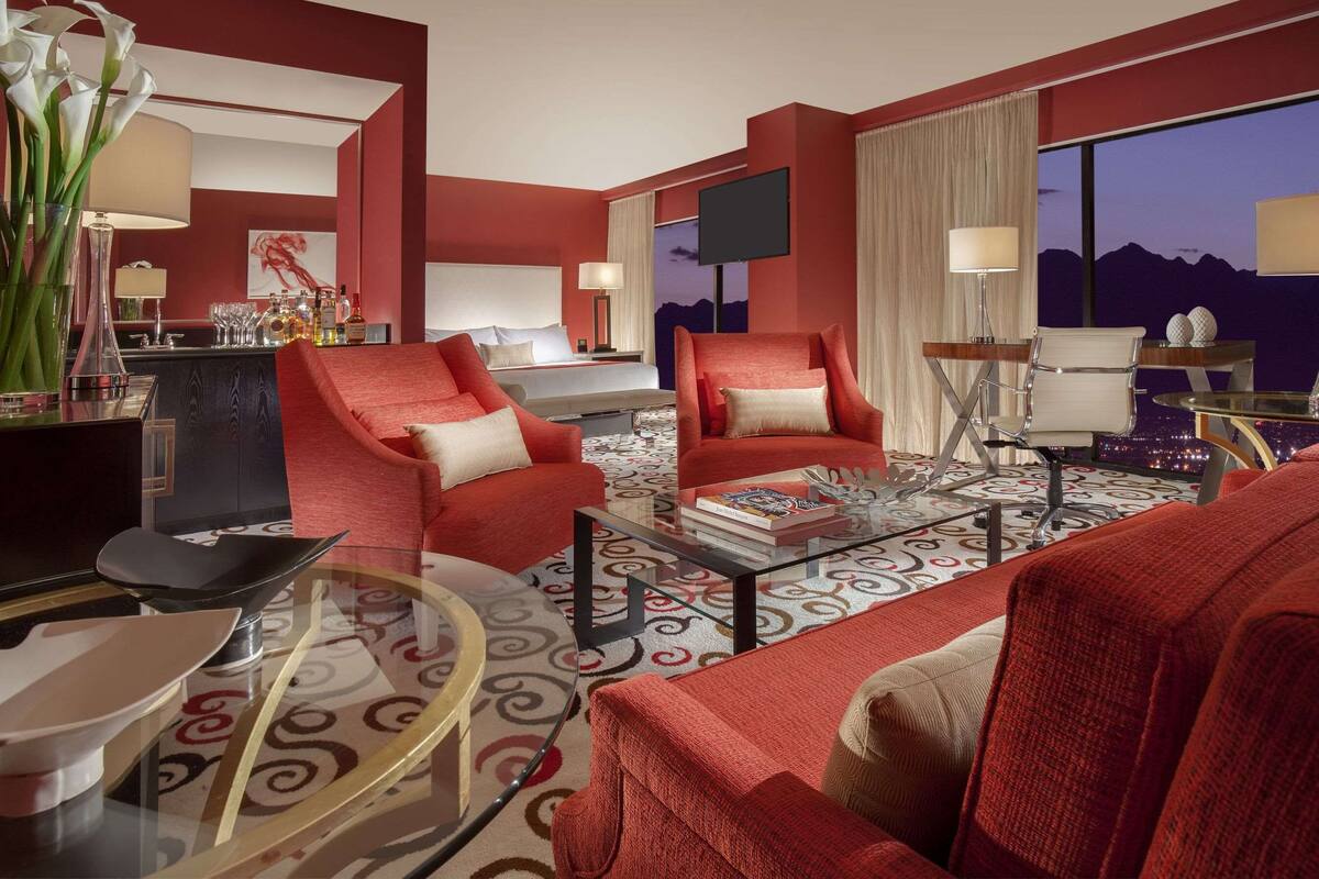Quarto de hotel moderno com decoração em vermelho e bege, uma mesa de centro redonda, uma televisão e vista para as montanhas ao entardecer.