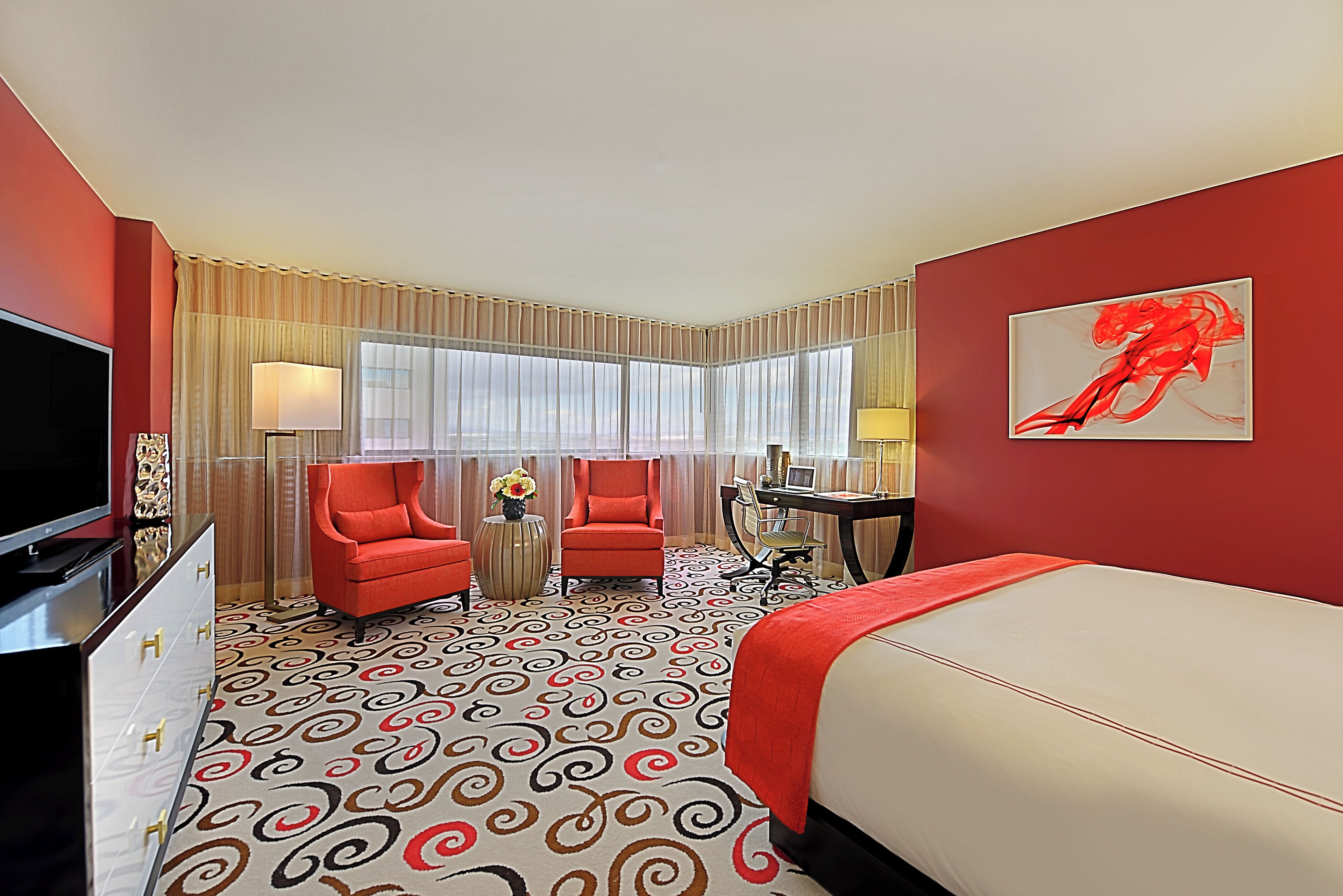 Ein Hotelzimmer mit einem großen Bett, zwei roten Sesseln, einem Schreibtisch mit Stuhl, einem Fernseher und einem großen Fenster mit Vorhängen.