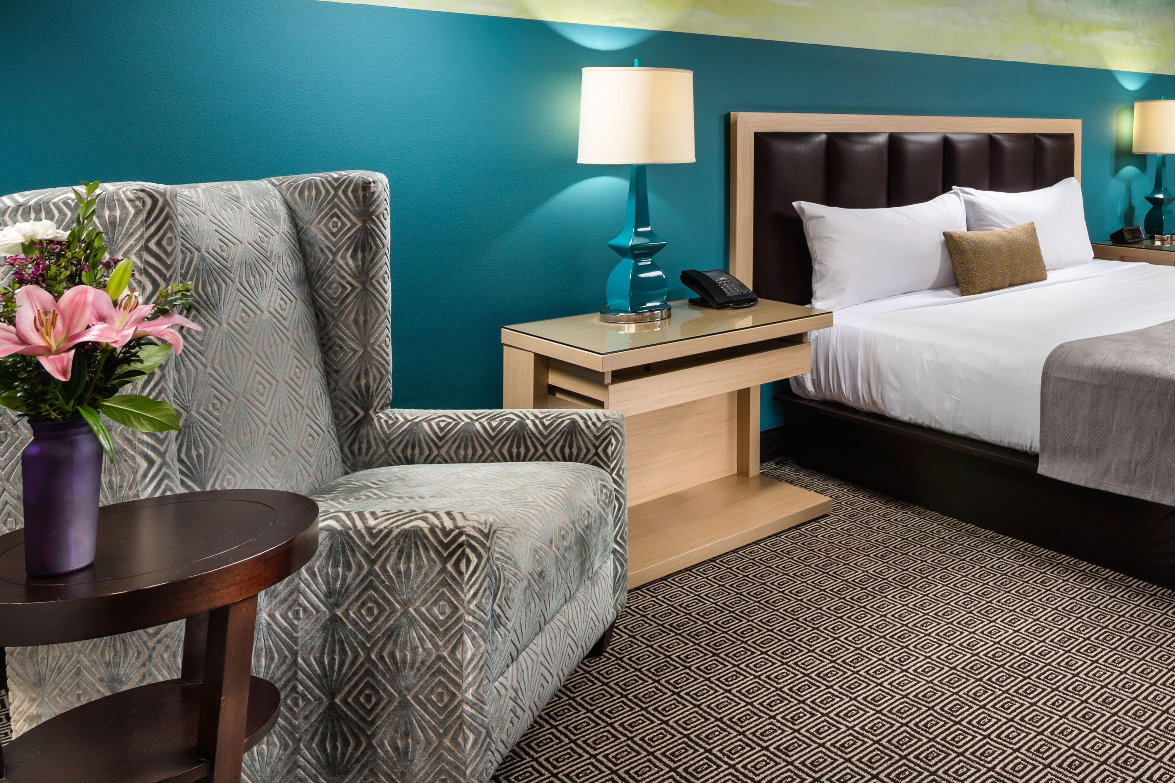 A hotel room with a bed, a chair, a nightstand with a lamp, and a vase with flowers.