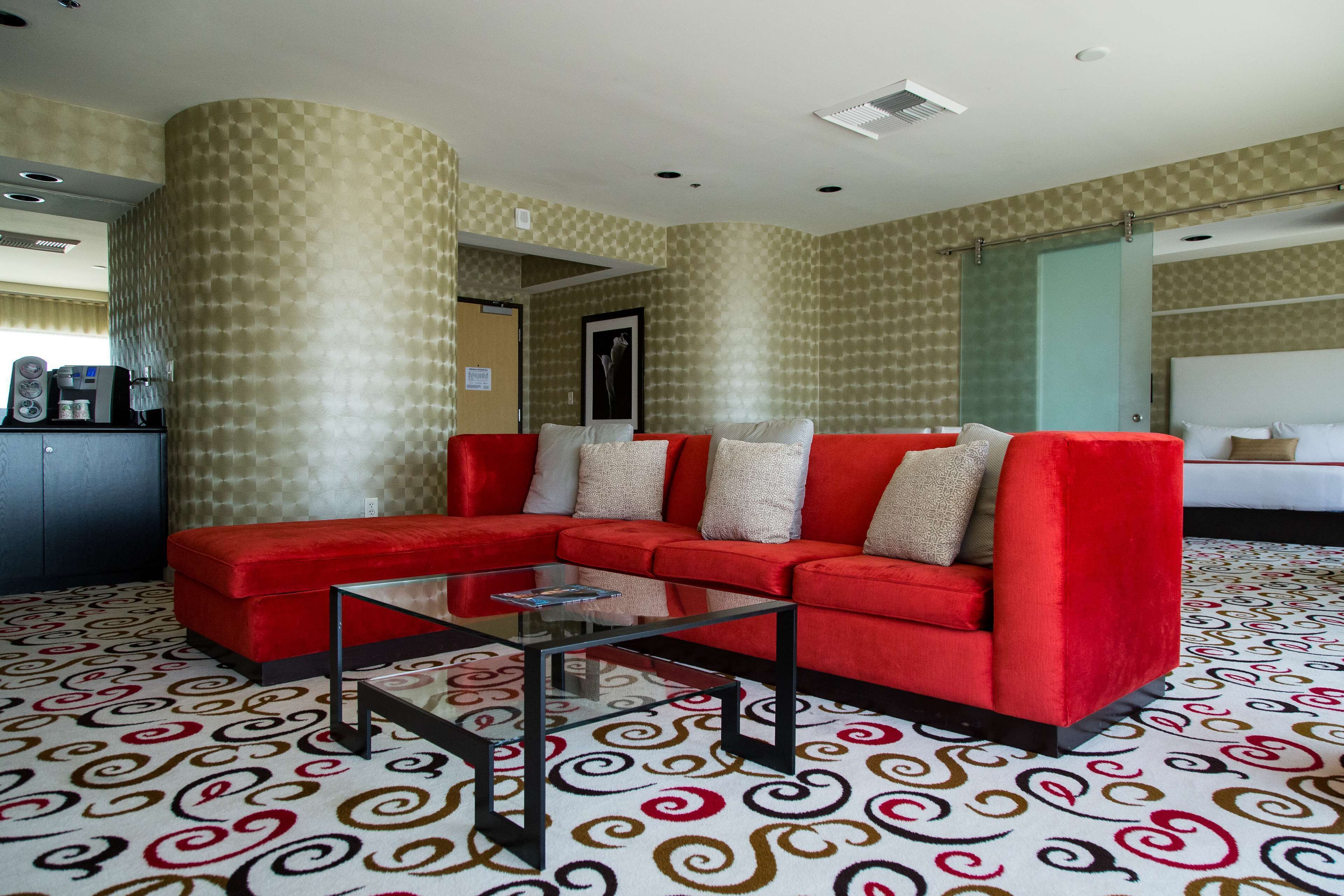 Un canapé d’angle rouge, une table basse en verre, une moquette à motifs et une porte donnant sur une autre pièce.