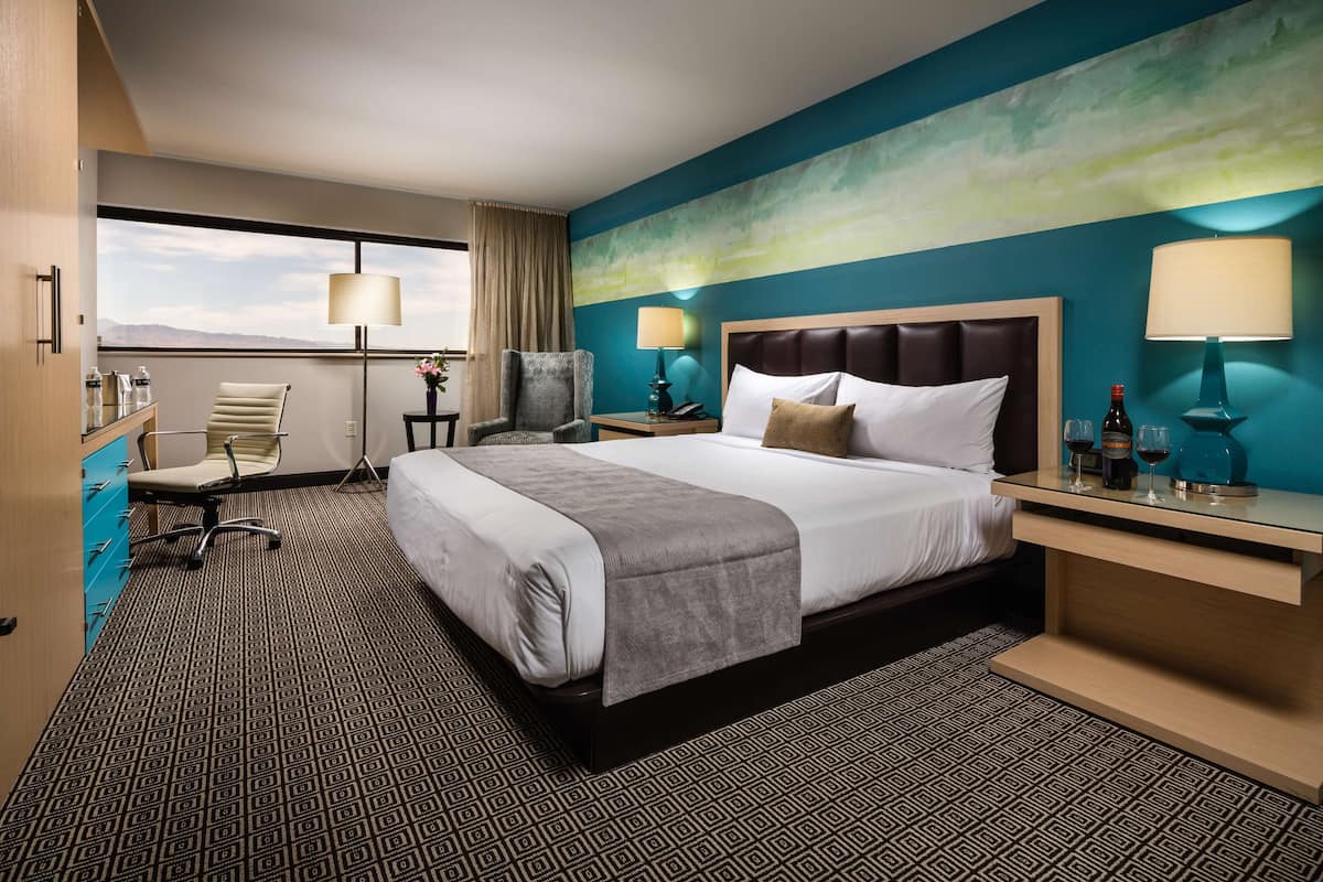 Quarto de hotel com uma cama grande, duas mesas de cabeceira, uma escrivaninha, uma cadeira, uma luminária de mesa, uma luminária de chão, uma janela com cortinas e vista para as montanhas.