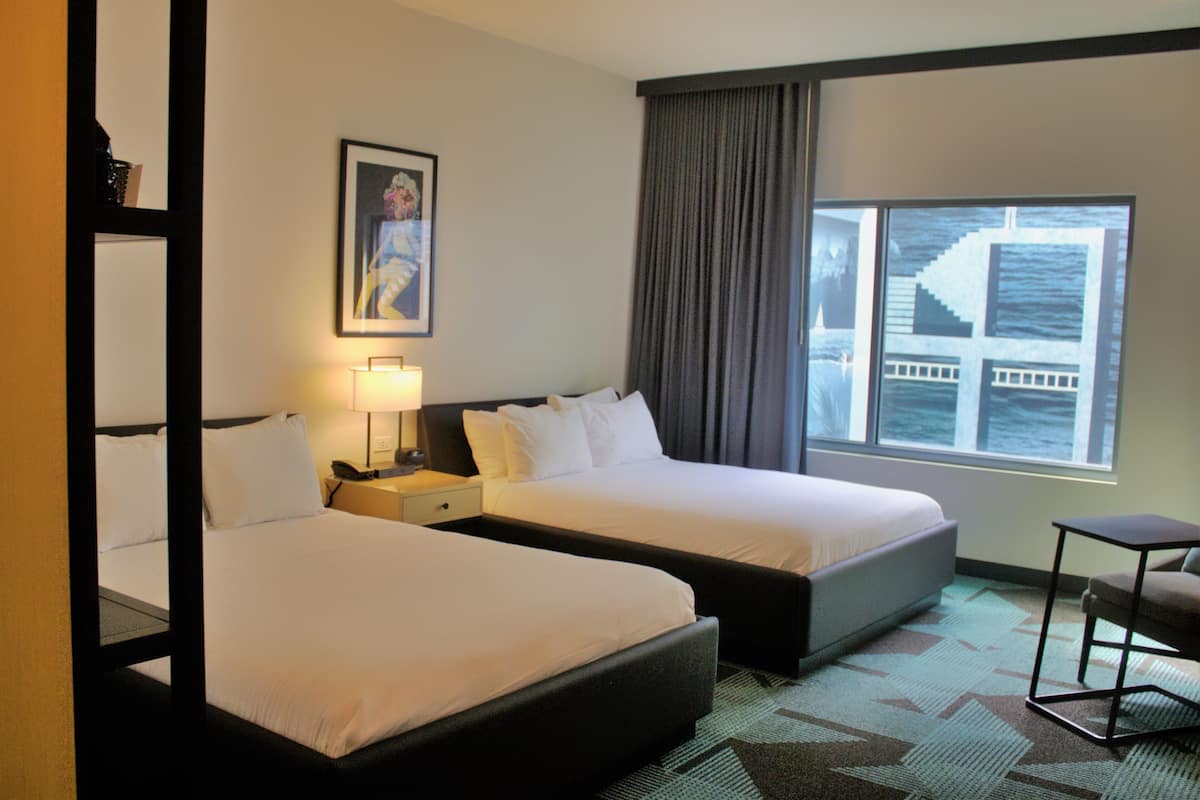 Quarto de hotel com duas camas, uma mesa de cabeceira com abajur, uma cadeira e uma janela grande com vista para um edifício.