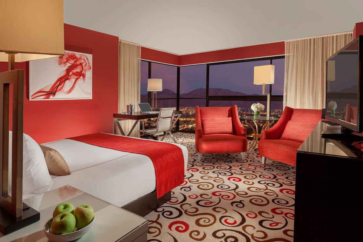 Quarto de hotel com uma cama grande, duas poltronas vermelhas, uma escrivaninha com dois laptops, uma janela ampla com vista para a cidade e um quadro abstrato vermelho na parede.