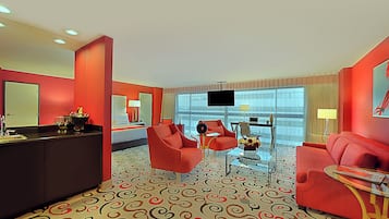 Habitación de hotel con alfombra de diseño rojo y beige, un televisor de pantalla plana grande, un sofá, una mesa de centro y una pequeña zona de bar.