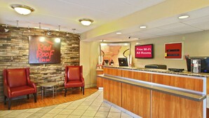 A Red Roof Inn reception area with a stone wall, two leather chairs, a wooden counter, and a sign indicating free Wi-Fi in all rooms. - Red Roof Inn Greensboro Coliseum (Greensboro)
