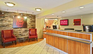 A Red Roof Inn reception area with a stone wall, two leather chairs, a wooden counter, and a sign indicating free Wi-Fi in all rooms. - Red Roof Inn Greensboro Coliseum (Greensboro)