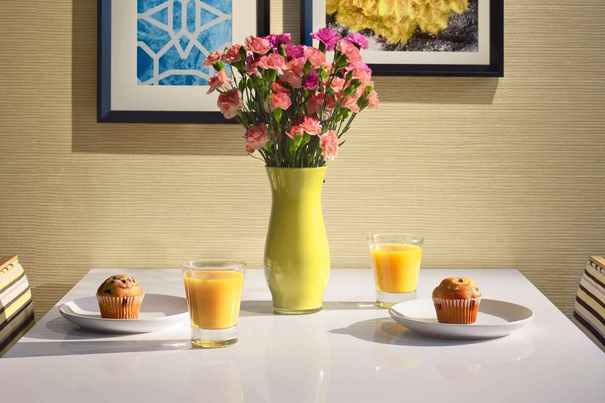 A table with two muffins, a vase with pink flowers, and two glasses of orange juice against a wall with framed artwork.