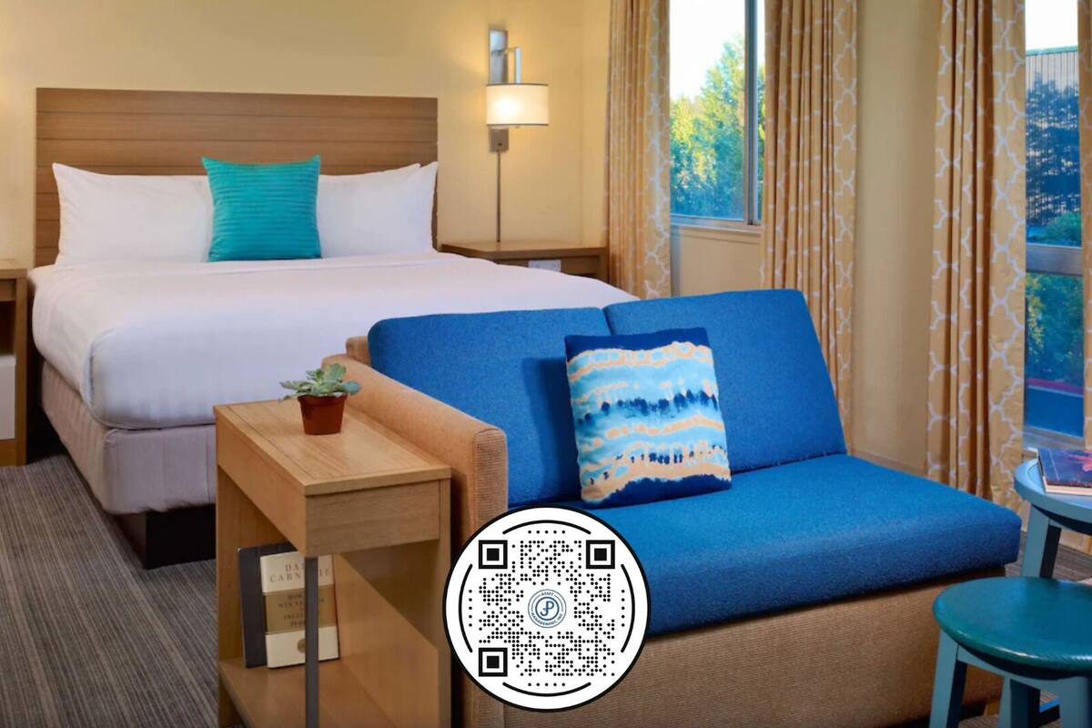 A hotel room with a bed, a sofa, a small table with a plant, and a window with curtains.