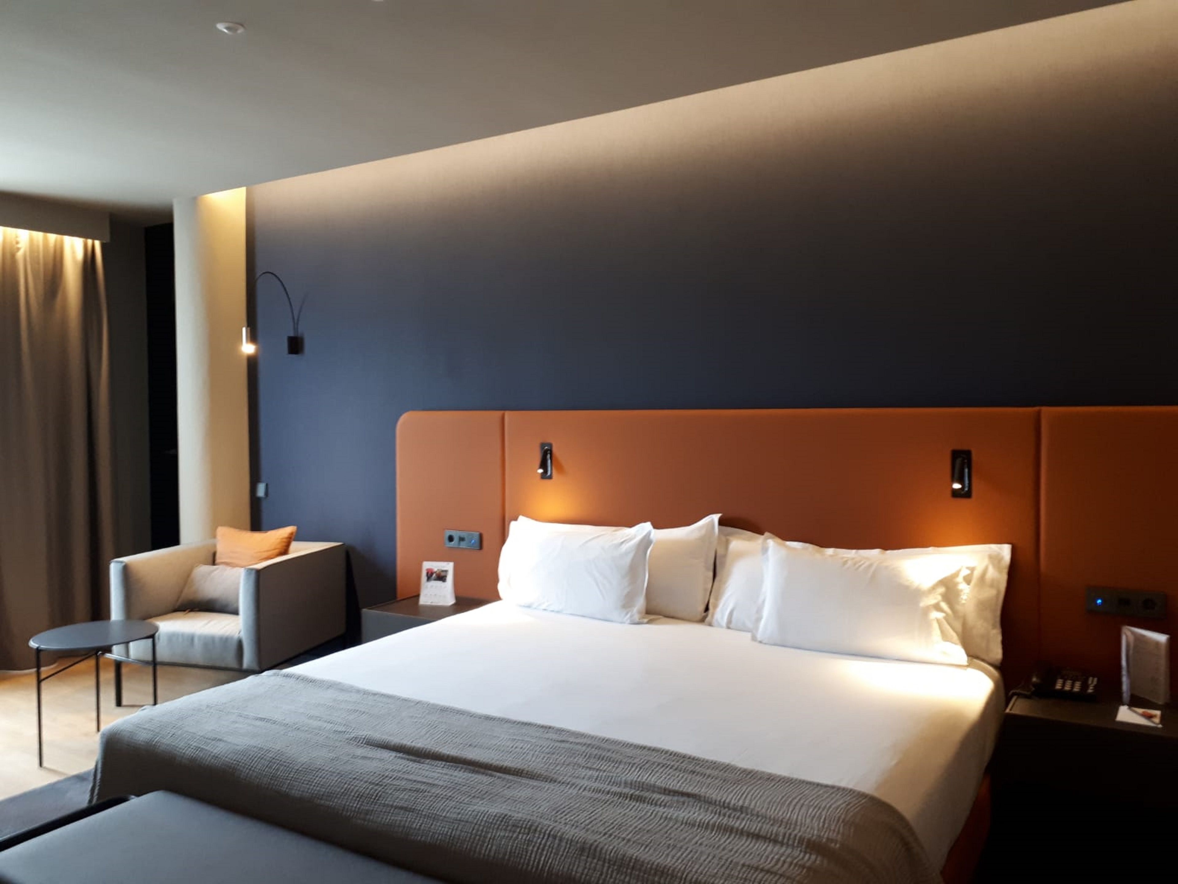 A modern hotel room with a large bed, a small sofa, a side table, and a desk.