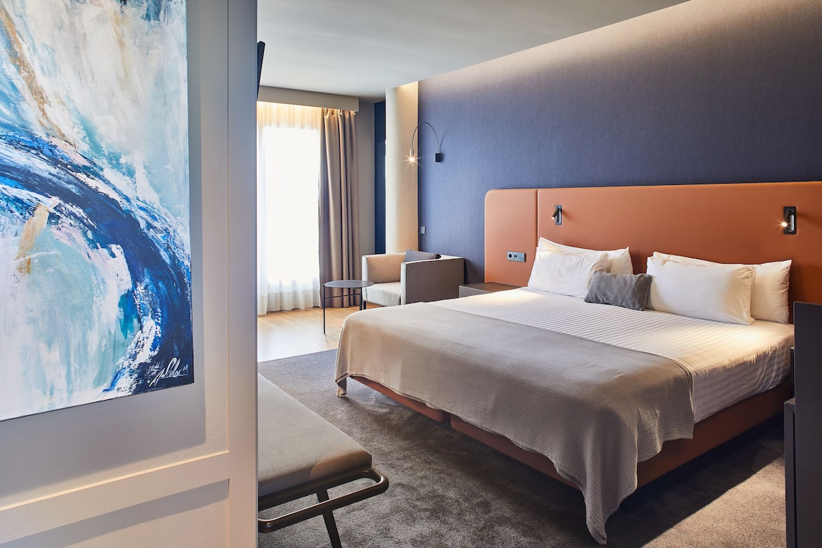 A modern hotel room with a large bed, a bedside table, a chair, and a view of the outdoors.