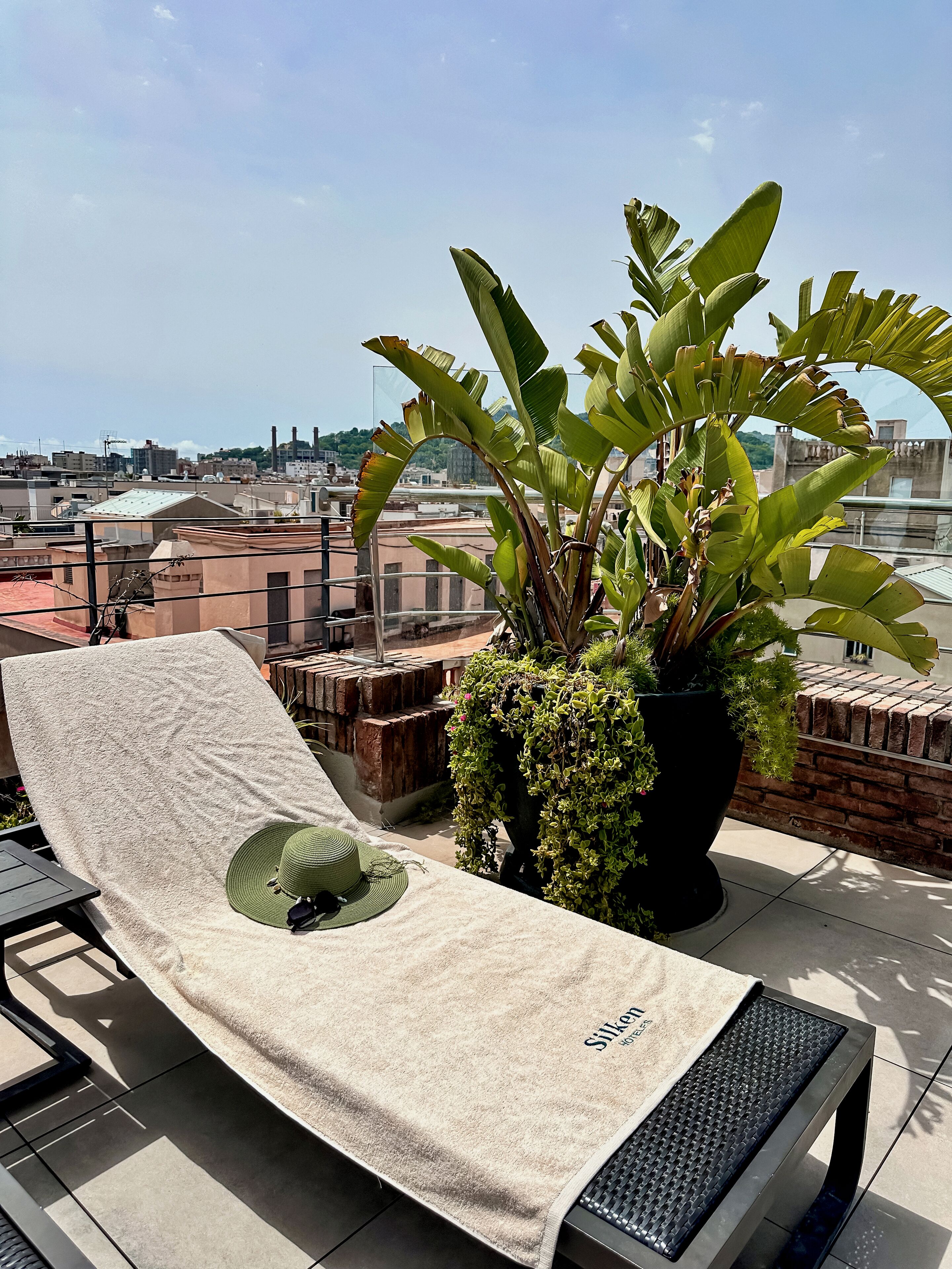 A rooftop lounge chair with a towel, a potted plant, and a view of the cityscape.