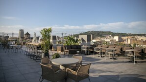 An outdoor patio with round tables and wicker chairs, overlooking a cityscape with buildings and hills. - Hotel Silken Ramblas (Barcelona)