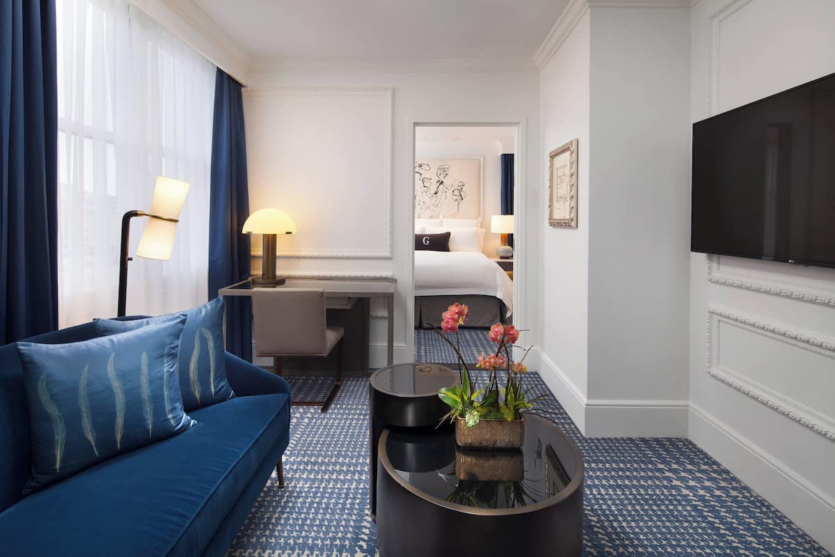 A hotel room with a blue sofa, a desk with a lamp, a bed with a nightstand, a flat-screen TV, and a patterned carpet.