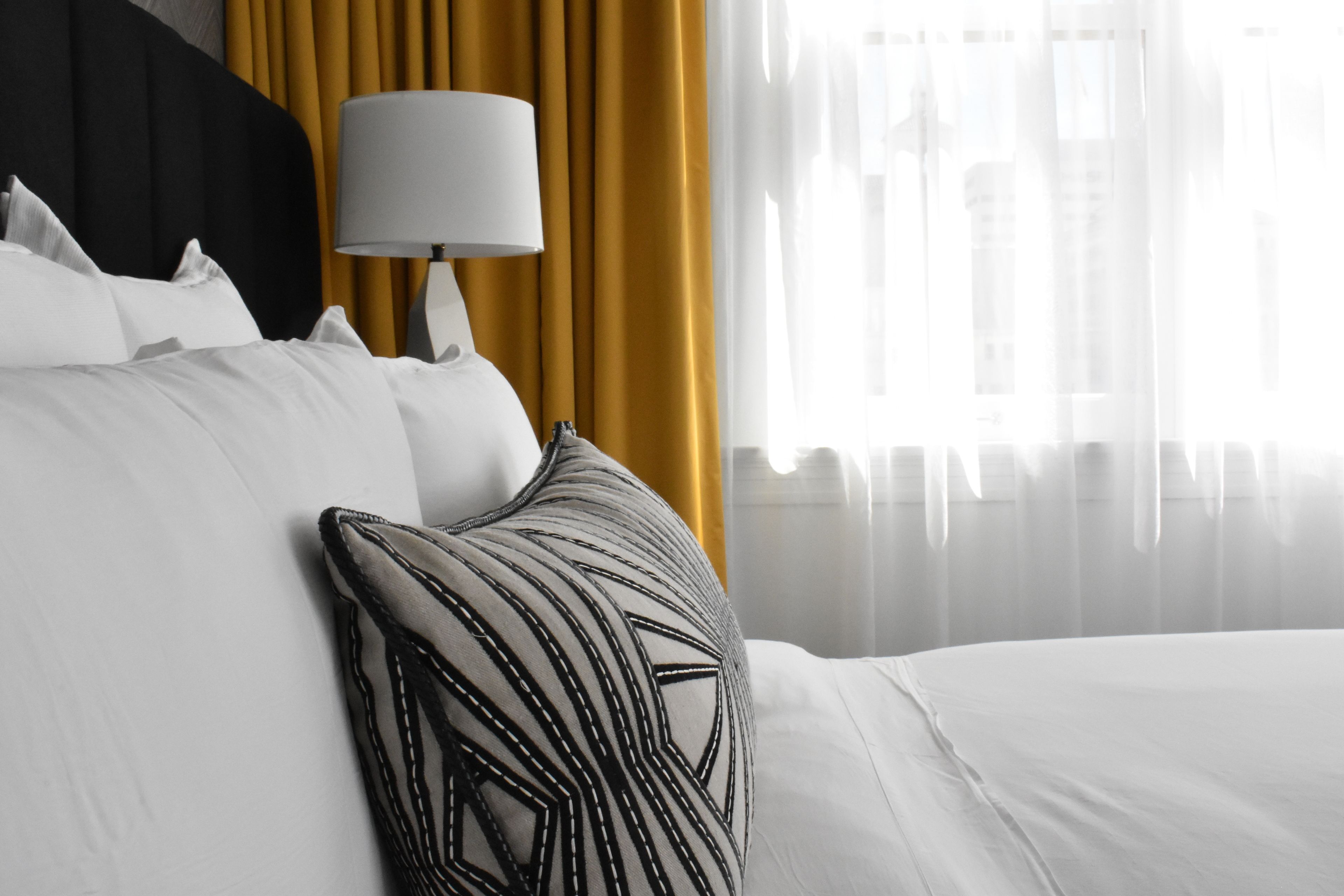 Una cama bien tendida con sábanas blancas y un cojín estampado con motivos geométricos, una lámpara de noche y una ventana con cortinas de gasa.