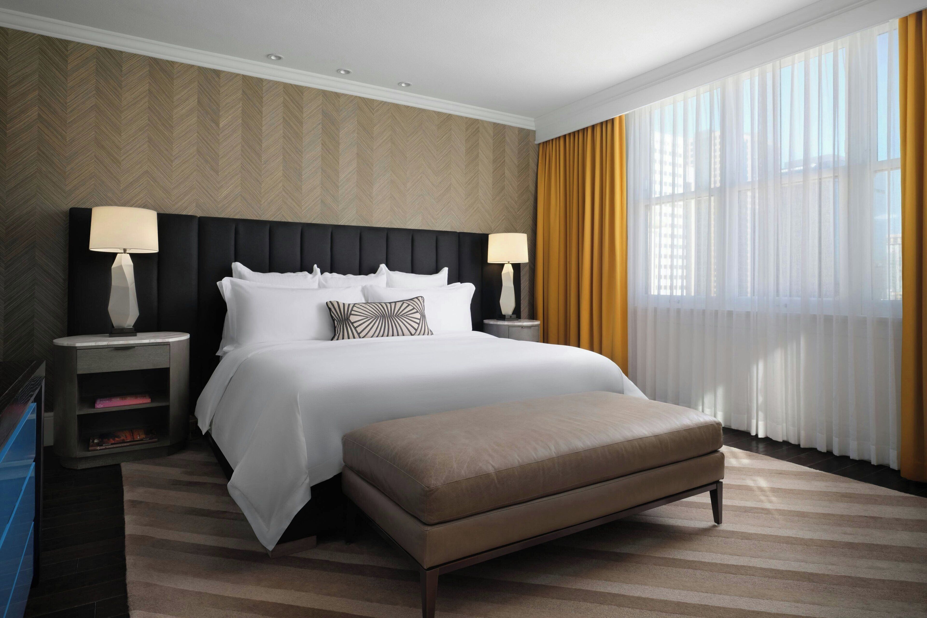 A hotel room with a large bed, two bedside tables with lamps, a bench, and a view of the city through sheer curtains.