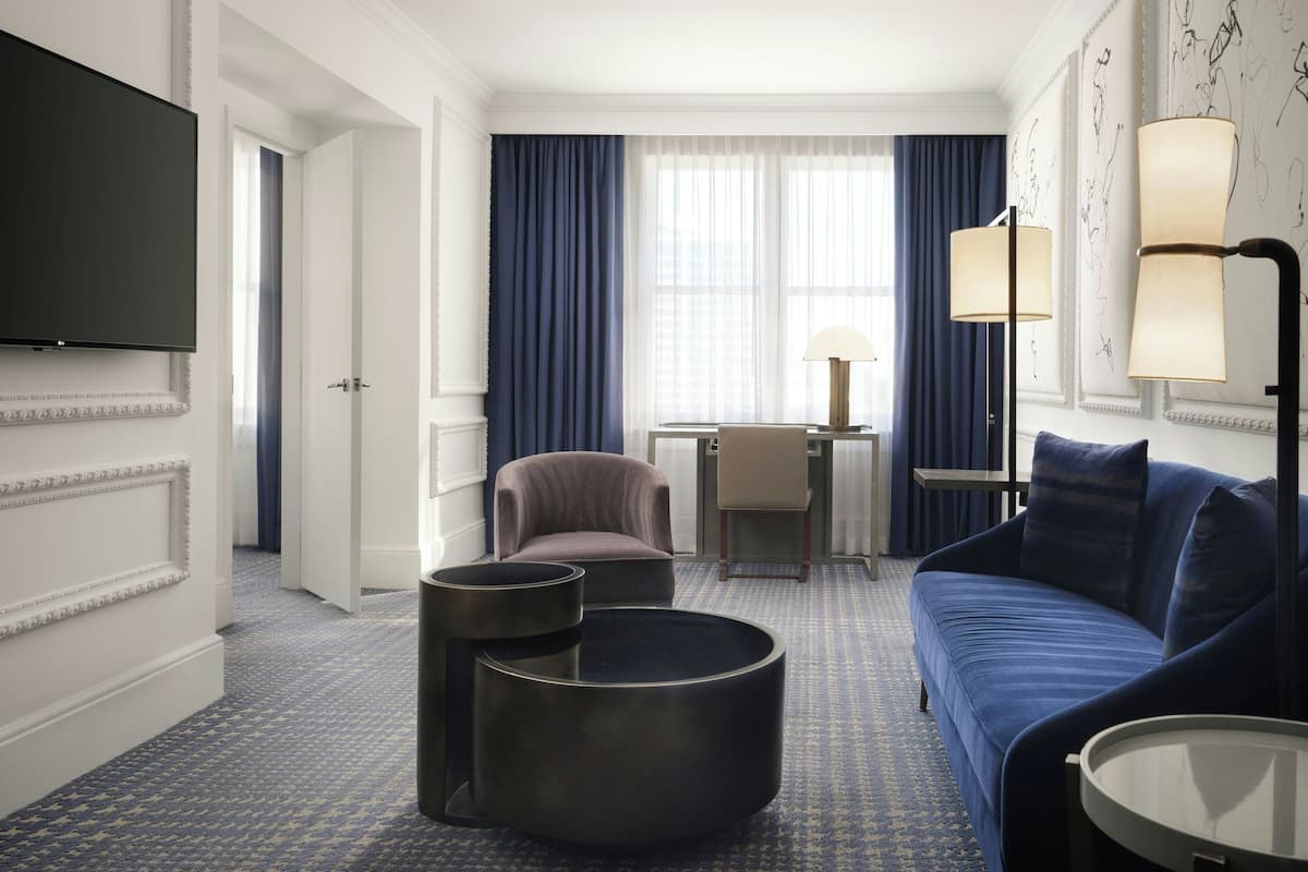 A hotel room with a blue sofa, a round coffee table, a TV, a desk with a chair, and a lamp.
