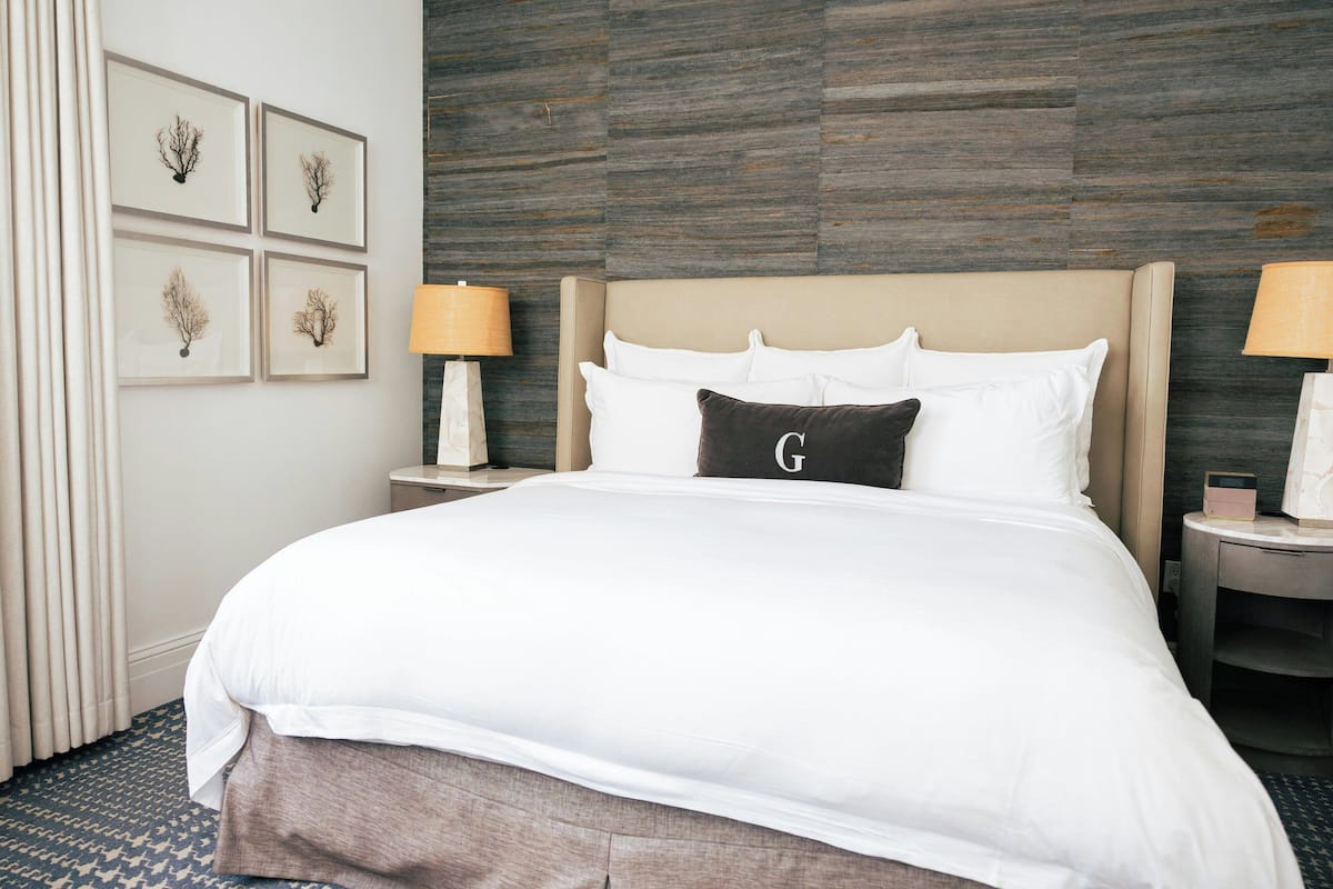 A neatly made bed with white linens and a single pillow with a 'G' on it. There are two bedside tables with lamps, and a wall with four framed pictures.
