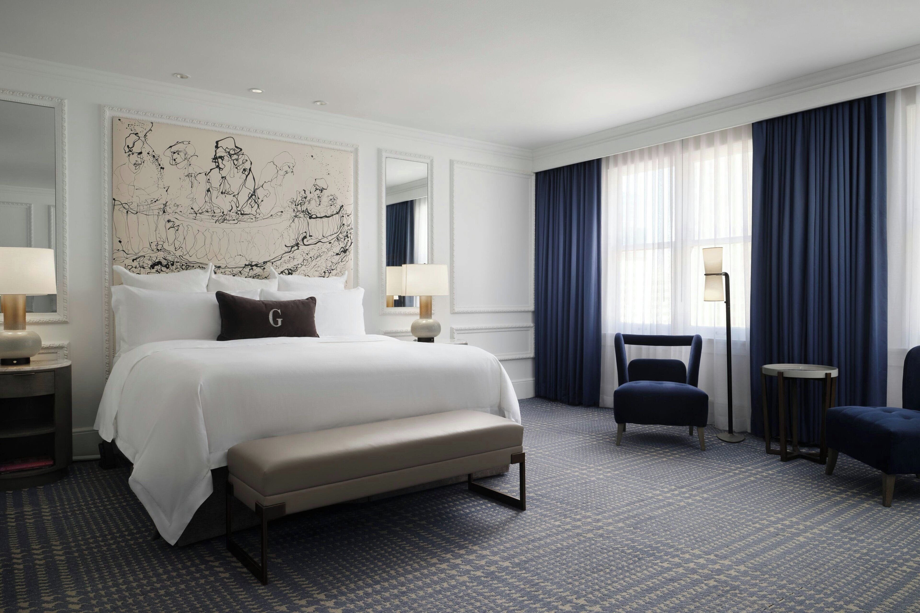 A hotel room with a large bed, a bench, two chairs, a desk, and a large abstract painting on the wall.