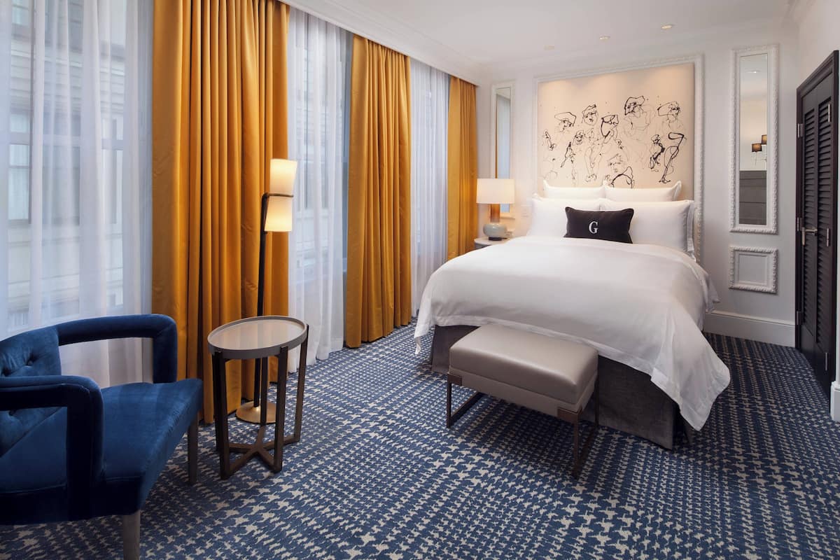 A hotel room with a large bed, a blue armchair, a small round table, and a view of the city through large windows with yellow curtains.