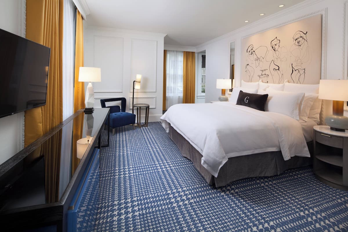 A hotel room with a large bed, a flat-screen TV, two bedside lamps, a desk with a chair, and a patterned carpet.