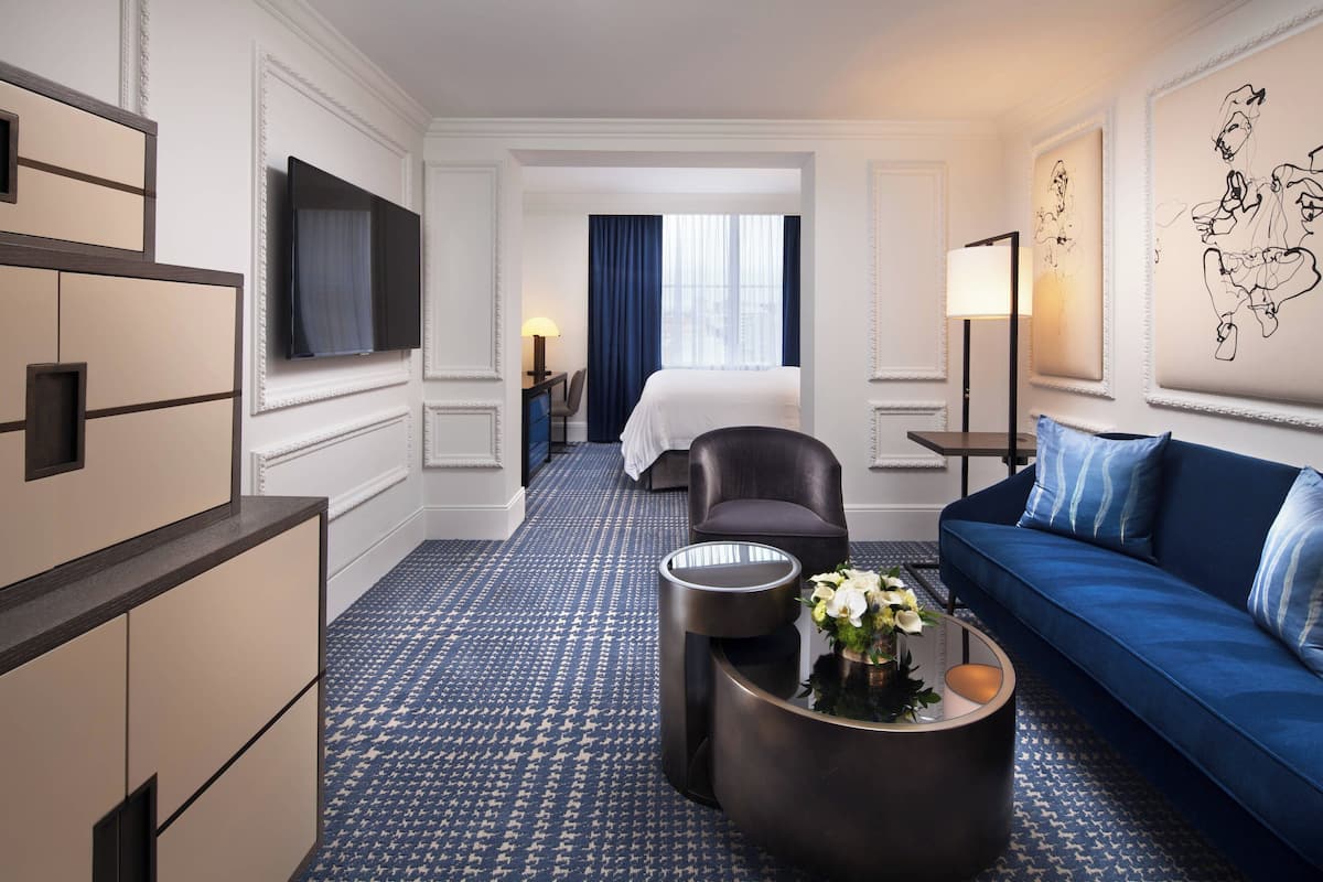 A hotel room with a large bed, a blue sofa, a coffee table with flowers, a TV mounted on the wall, and a patterned carpet.