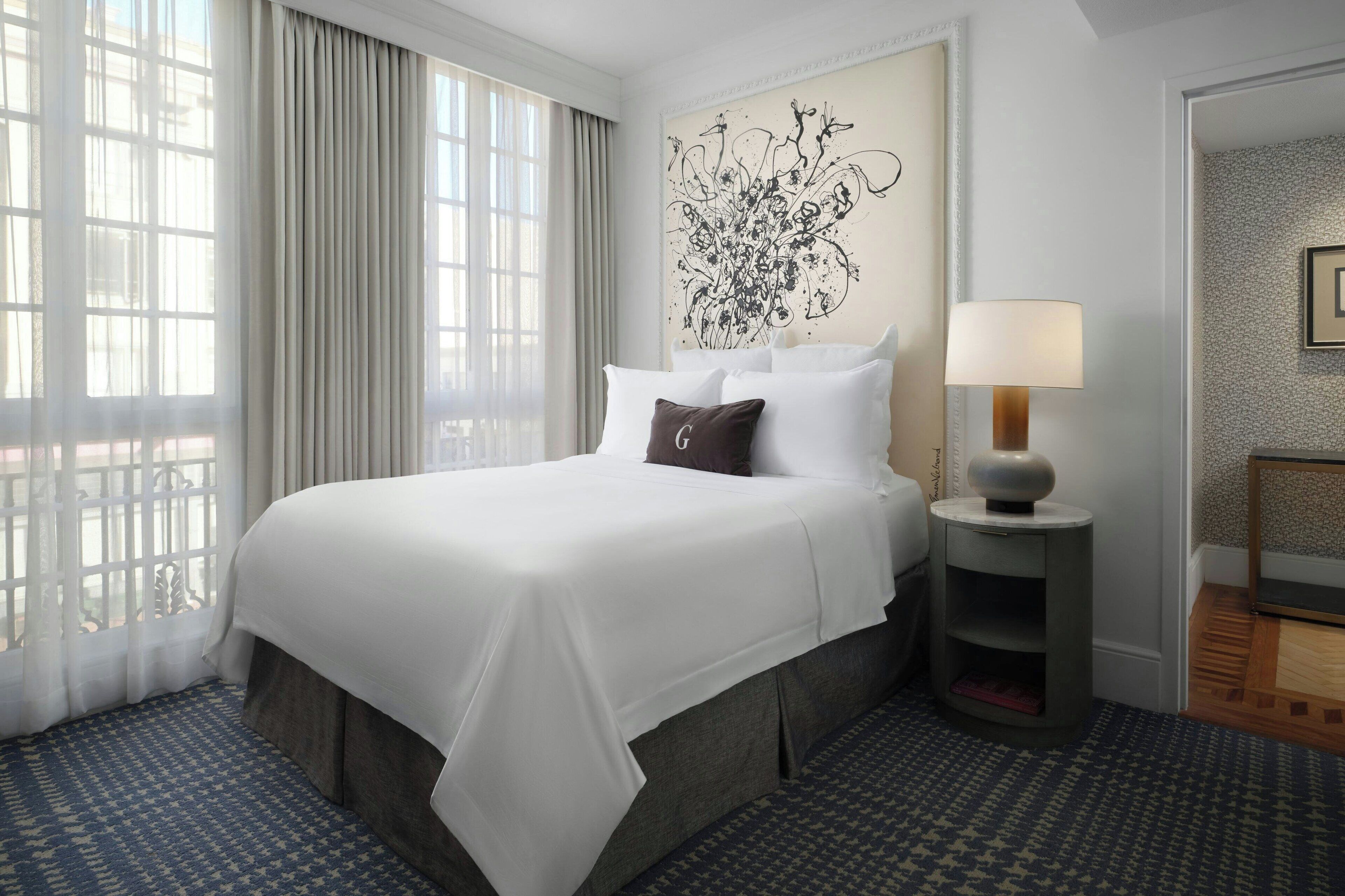 Ein Hotelzimmer mit einem großen Bett, einem Nachttisch mit Lampe, einem Fenster mit Vorhängen und einer Wand mit einer floralen Dekoration.