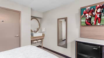 Una habitación de hotel con una cama, una kitchenette pequeña, un espejo y un televisor mostrando un partido deportivo.