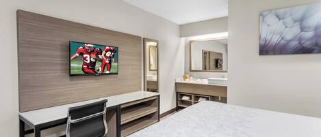 A hotel room with a bed, a desk with a chair, a television mounted on the wall, a bathroom with a mirror, and a painting on the wall.