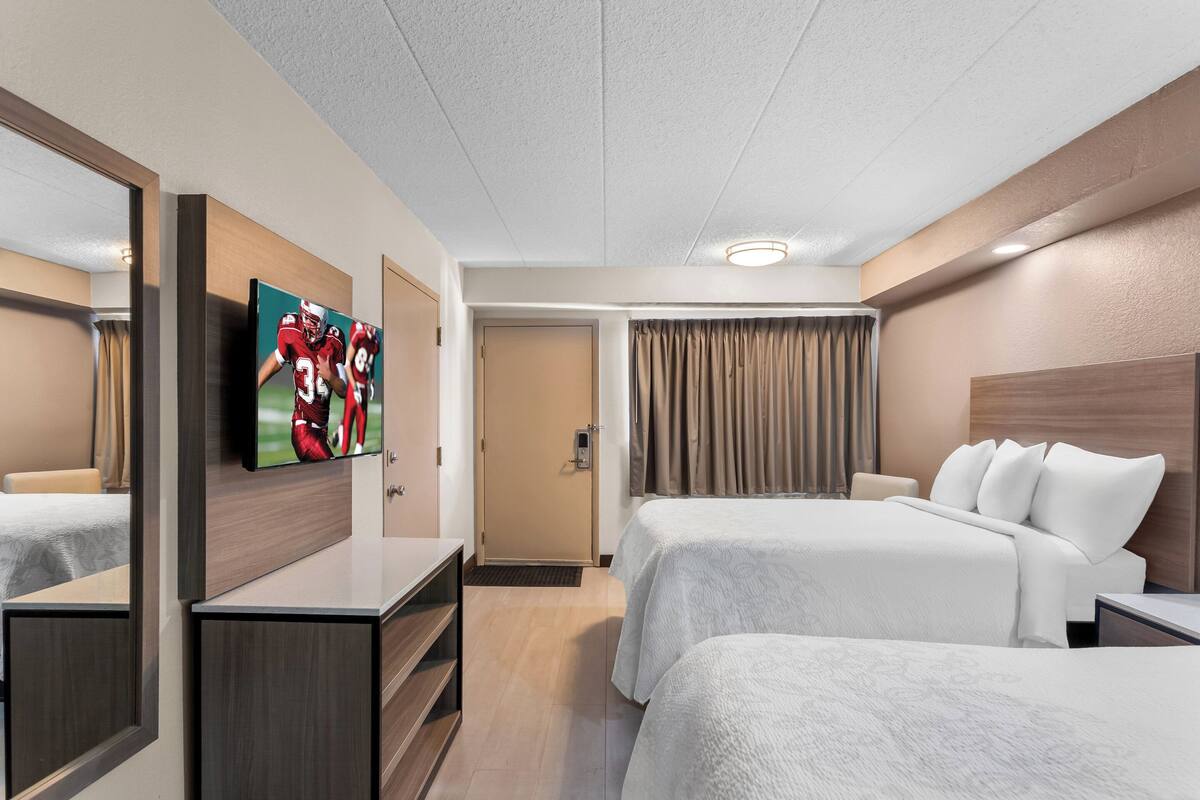 Ein Hotelzimmer mit zwei Betten, einem Fernseher, einem Schreibtisch und einem Spiegel.