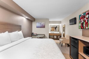 A hotel room with a large bed, a desk, a chair, a television, a refrigerator, and a bathroom with a mirror and sink. - Red Roof Inn PLUS+ Chicago - Hoffman Estates (Hoffman Estates)