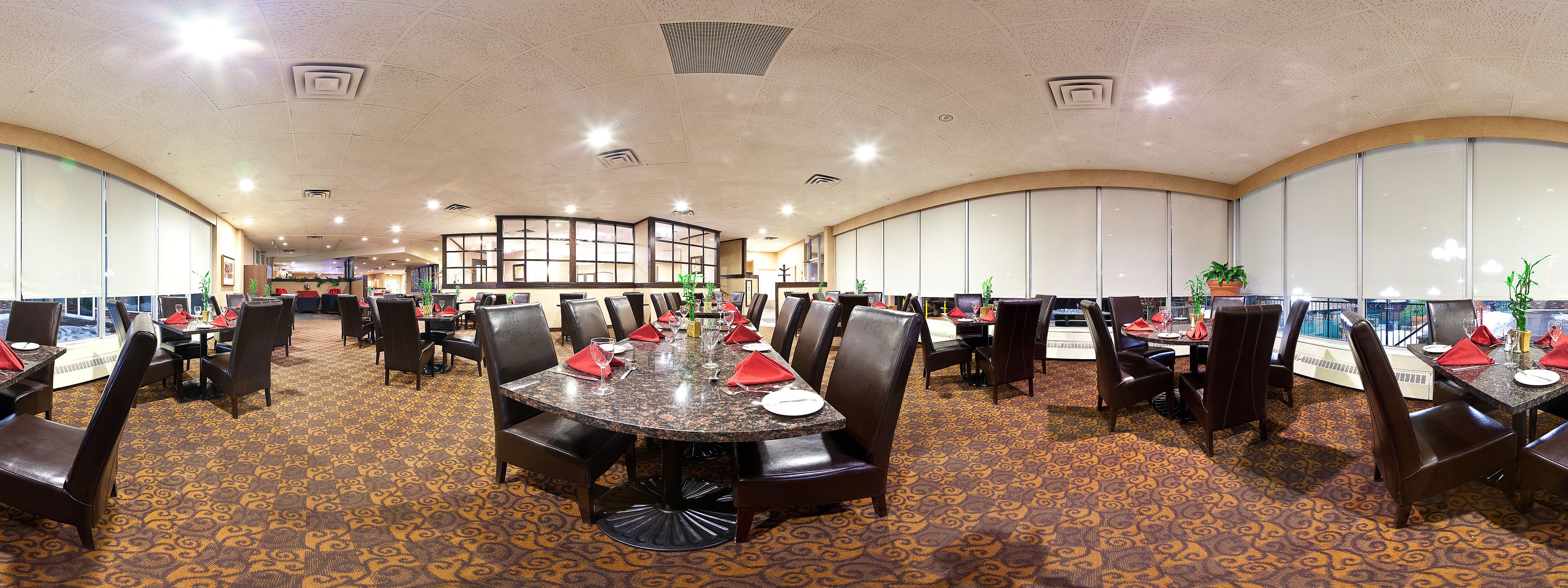 Un restaurante espacioso con techos altos, ventanas amplias y una alfombra estampada. Las mesas están dispuestas con servilletas rojas y copas.