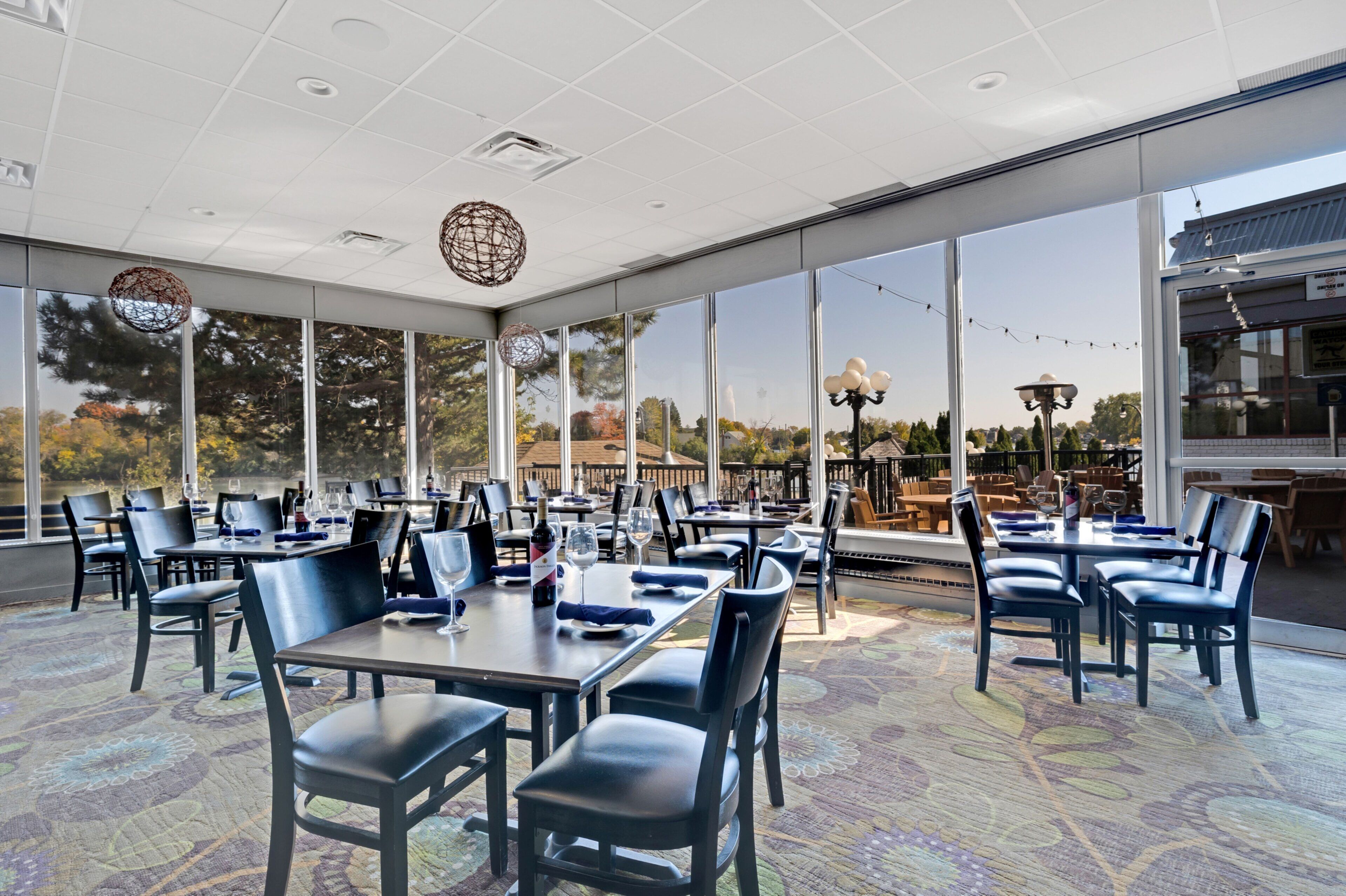 A spacious indoor dining area with large windows, tables set with chairs, and a view of trees and buildings outside.
