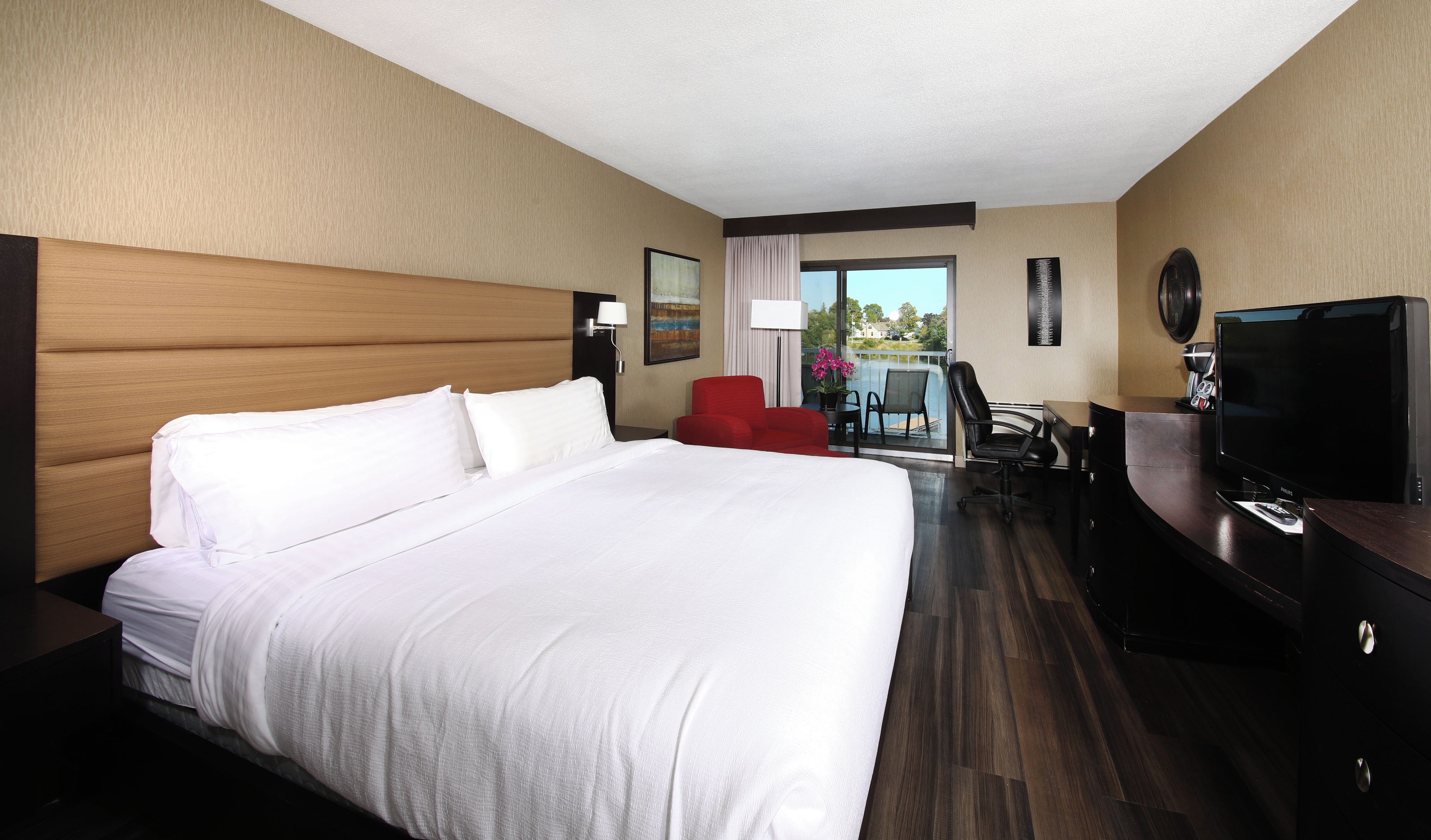 A hotel room with a large bed, a desk with a flat-screen TV, a red chair, and a view of the outdoors.
