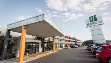 A Holiday Inn hotel with a covered entrance, a parking lot with several cars, and a clear sky.